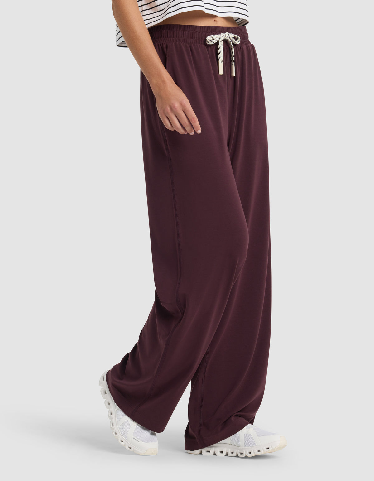 A person is shown from the waist down wearing Cozy Earth Women's Studio Wide Leg Pant in burgundy with a drawstring waist, paired with white sneakers and a cropped striped top.