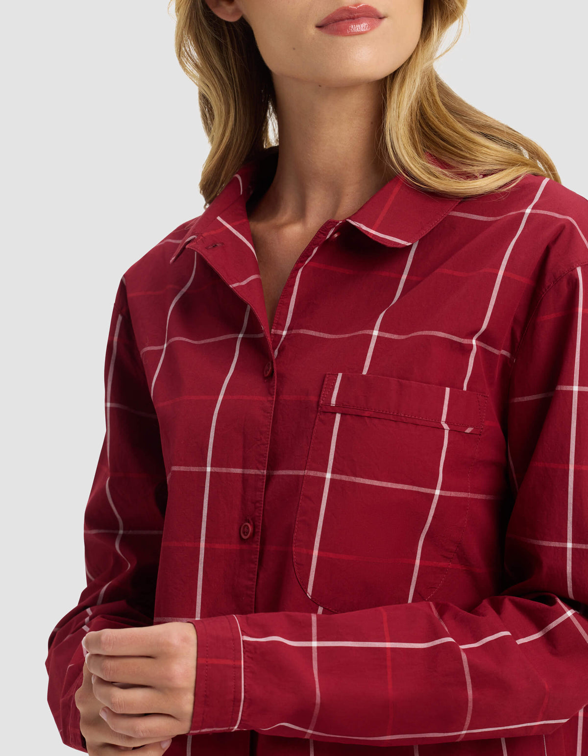 A woman wears the Cozy Earth Women's Sutton Long Sleeve Pajama Top, a red plaid button-up shirt with white lines. She stands against a plain background, shown from the shoulders down with her hands gently clasped together.