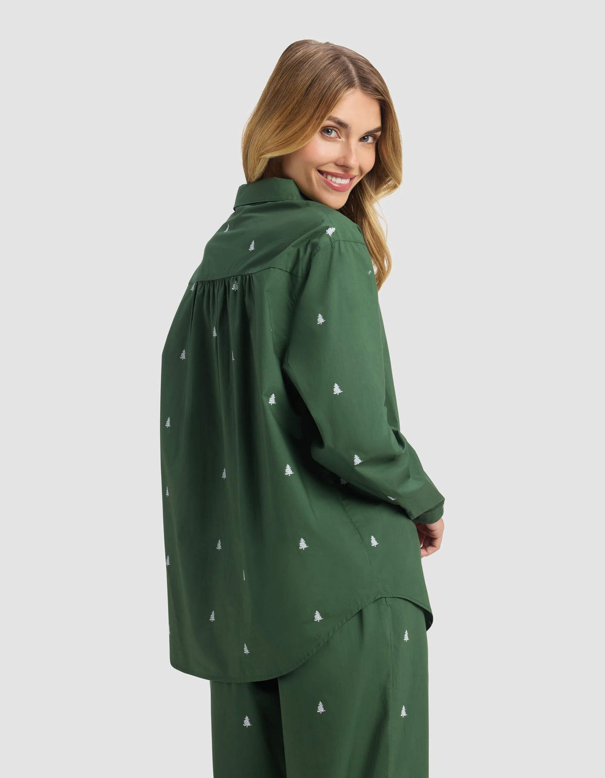 A woman with long blonde hair smiles over her shoulder, wearing the Cozy Earth Women's Sutton Long Sleeve Pajama Top in dark green, paired with matching pants featuring small white embroidered tree patterns. The background is light gray.