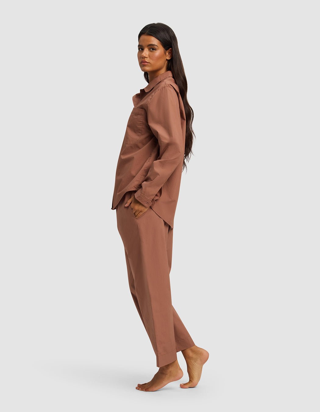 A woman with long dark hair stands barefoot in profile, wearing the Cozy Earth Women's Sutton Long Sleeve Pajama Set in light brown, her hands in her pockets against a plain light background.|Color:Saddle