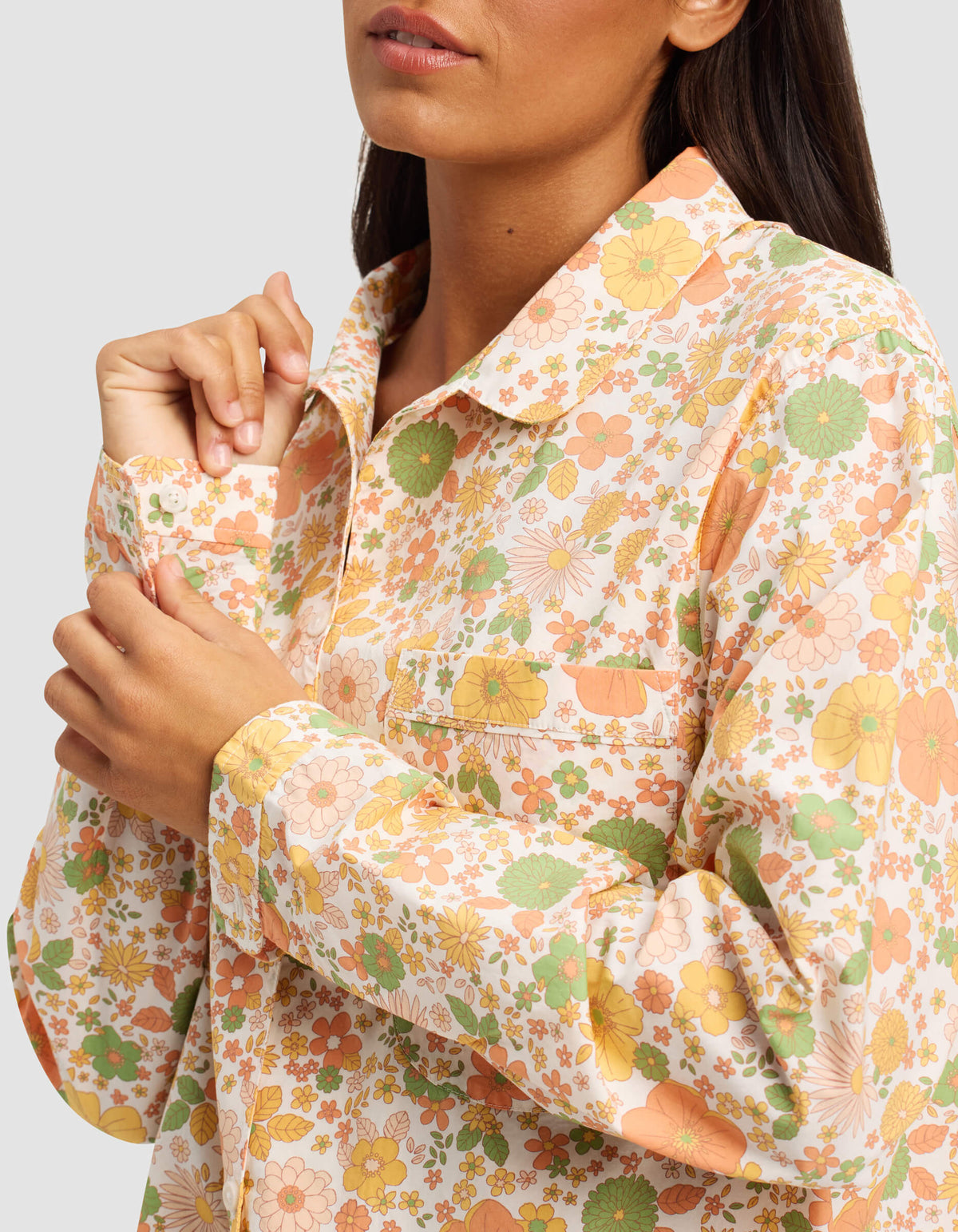 A woman buttons the collar of the Cozy Earth Women's Sutton Long Sleeve Pajama Top, featuring a colorful floral pattern in orange, green, and yellow. Her face is partially visible against a plain light gray background.