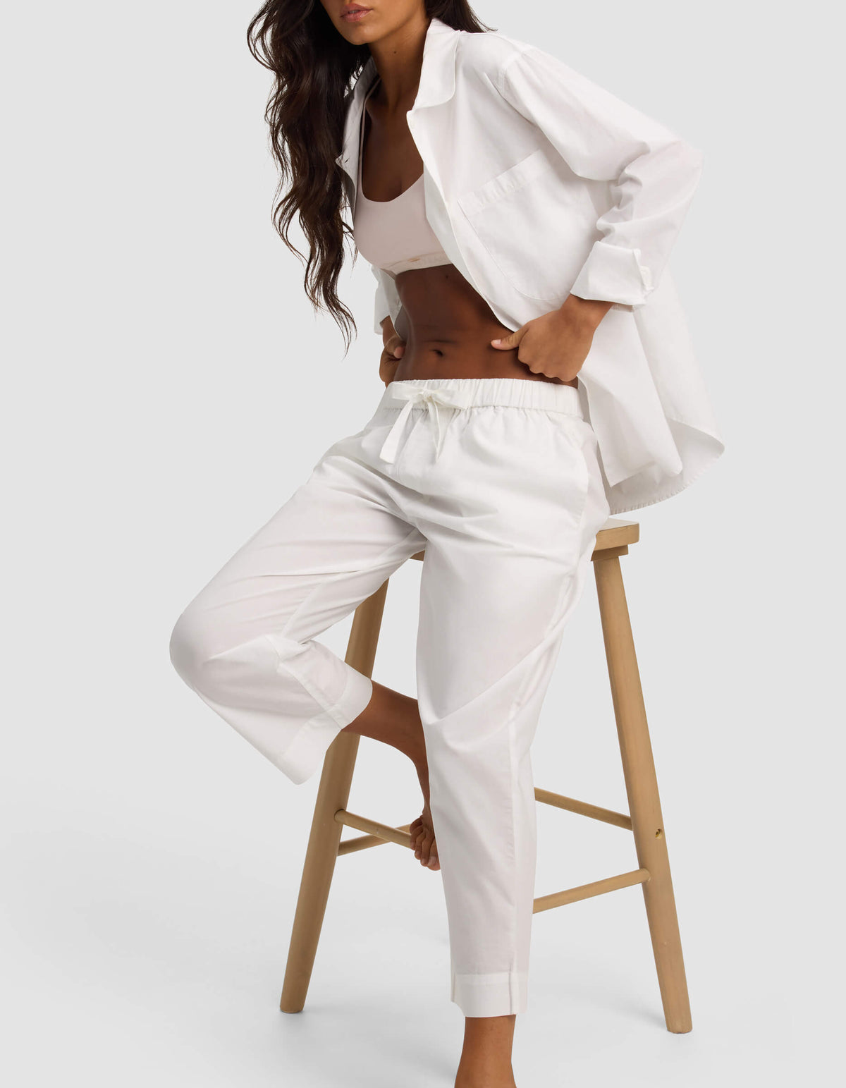 A woman with long wavy hair sits on a wooden stool against a plain light background, wearing Cozy Earth's Women's Sutton Cropped Pajama Pant paired with a white button-up shirt and crop top.