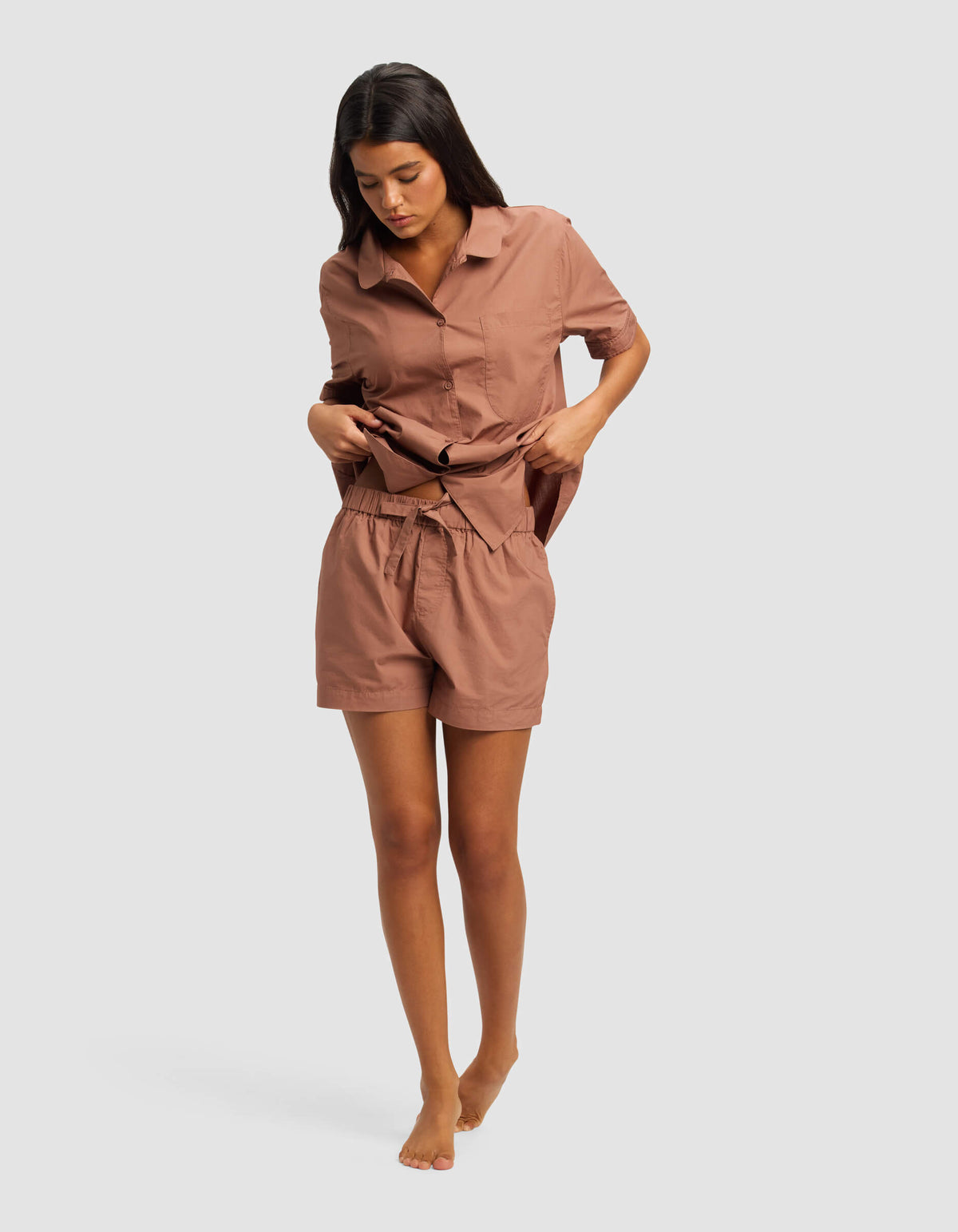 A woman wearing Cozy Earth's Women's Sutton Pajama Short with a matching loose, short-sleeved, button-up shirt stands barefoot against a plain light background, looking down and adjusting her shirt.