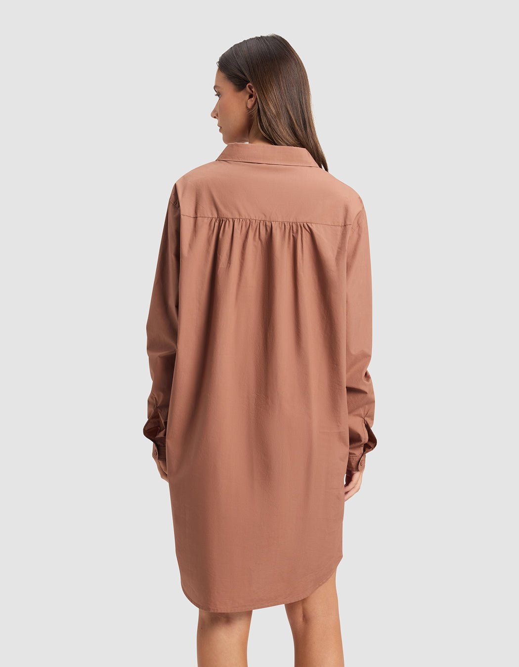 A woman faces away from the camera, wearing Cozy Earth's Women's Sutton Sleep Dress—a loose, long-sleeved, knee-length brown shirt dress with a collar—against a plain light gray background.