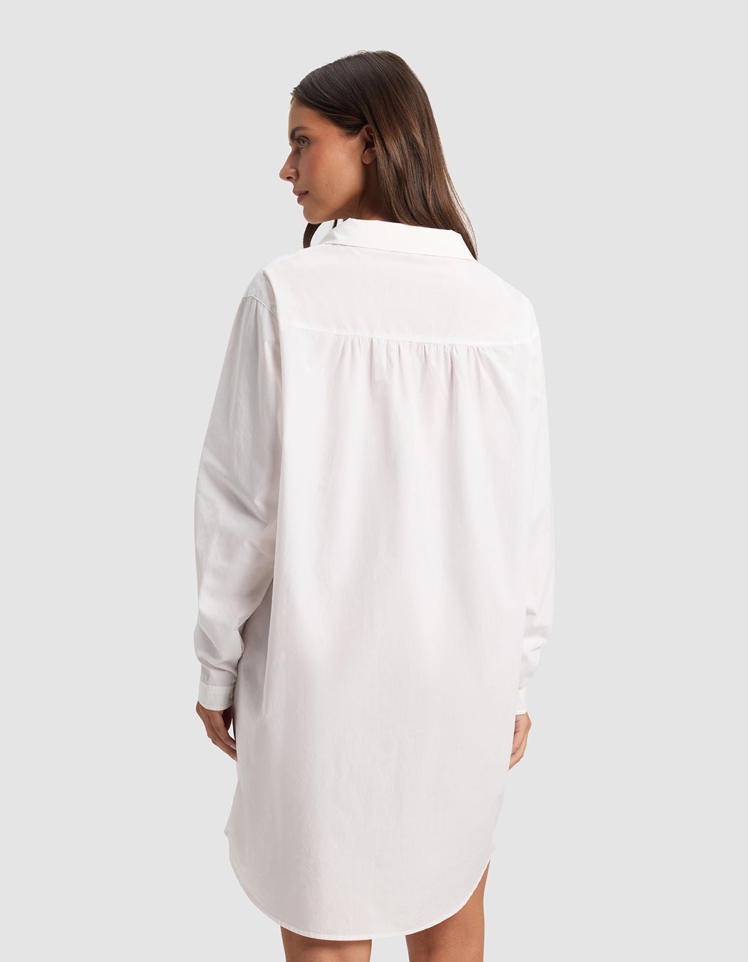 A woman with long brown hair faces away, wearing the Cozy Earth Women's Sutton Sleep Dress—a loose-fitting white, long-sleeve shirt dress—set against a plain light gray background.|Color:White