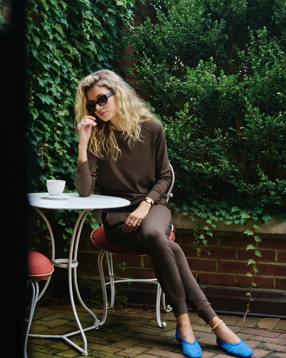 A woman with blonde hair and sunglasses sits at an outdoor patio table wearing a brown long-sleeve top and matching brown pants, with bright blue shoes, against a background of green ivy and brick. 