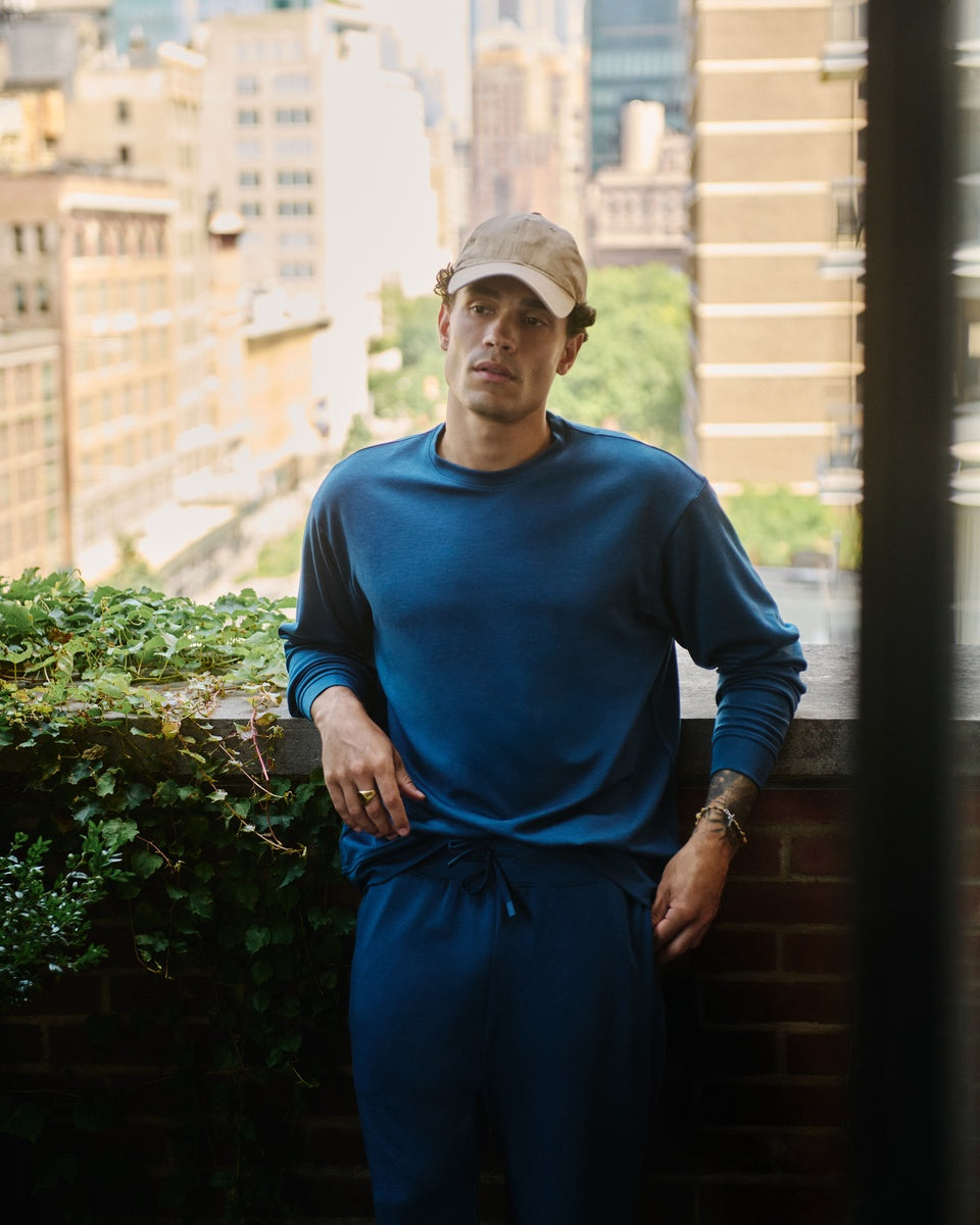 A man wearing a blue Cozy Earth Versa crewneck and matching blue Versa Joggers stands on a balcony with a city skyline background.