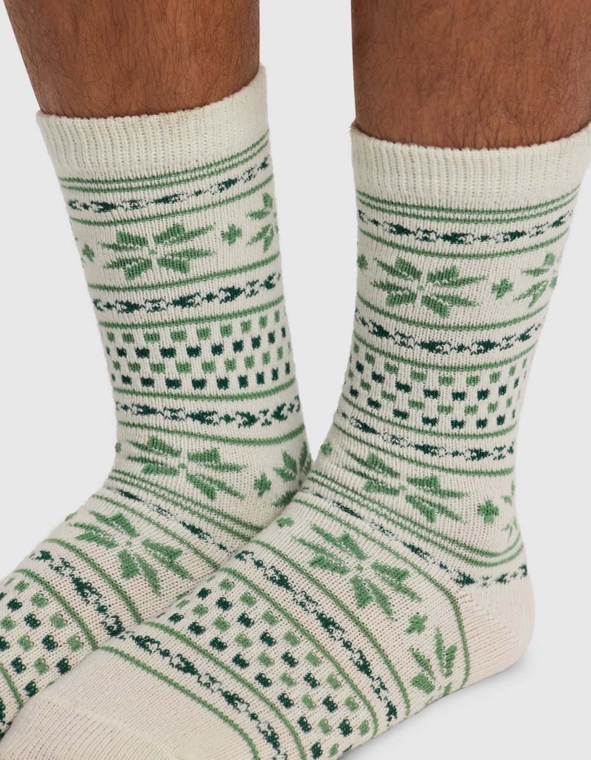 Close-up of a person’s lower legs wearing Cozy Earth’s The Snowflake Plush Lounge Sock in cream with green geometric and snowflake patterns against a plain white background.