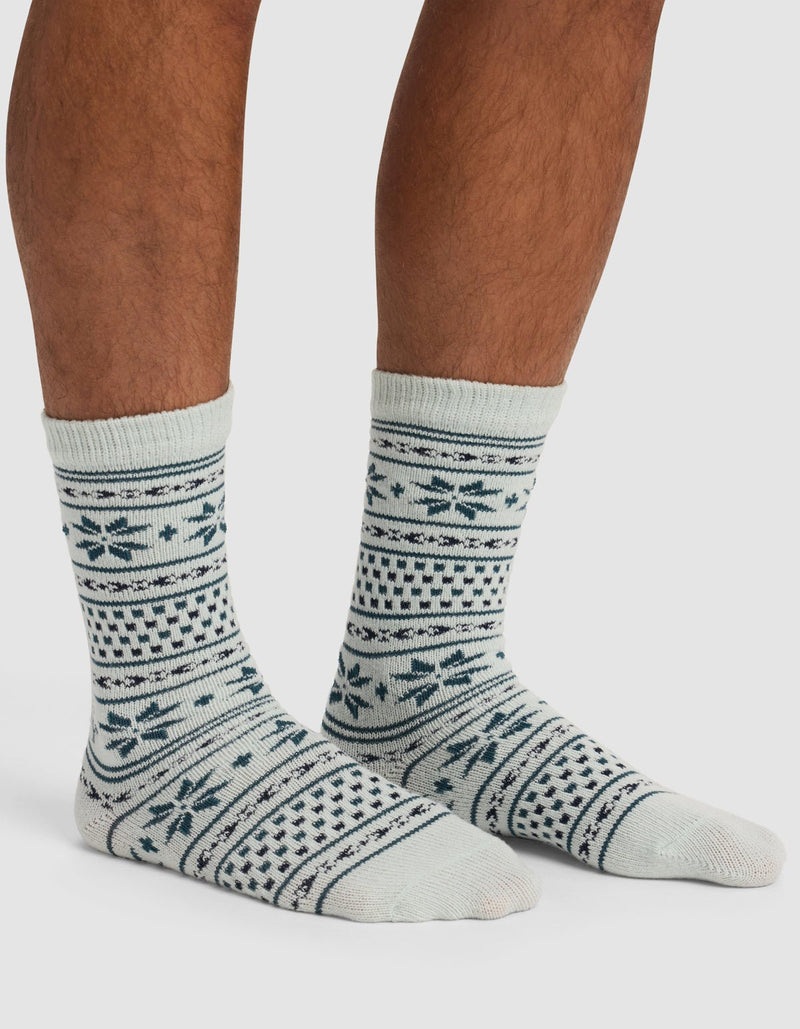 A person wearing Cozy Earth's The Snowflake Plush Lounge Sock—light-colored with dark blue geometric and snowflake patterns—stands on a light surface, showing only their lower legs and feet.