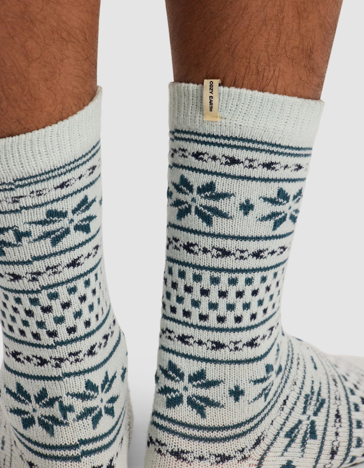 A close-up shows medium brown feet wearing Cozy Earth's The Snowflake Plush Lounge Sock—light-colored with blue Nordic-style patterns and a small beige tag.|Color:Mist