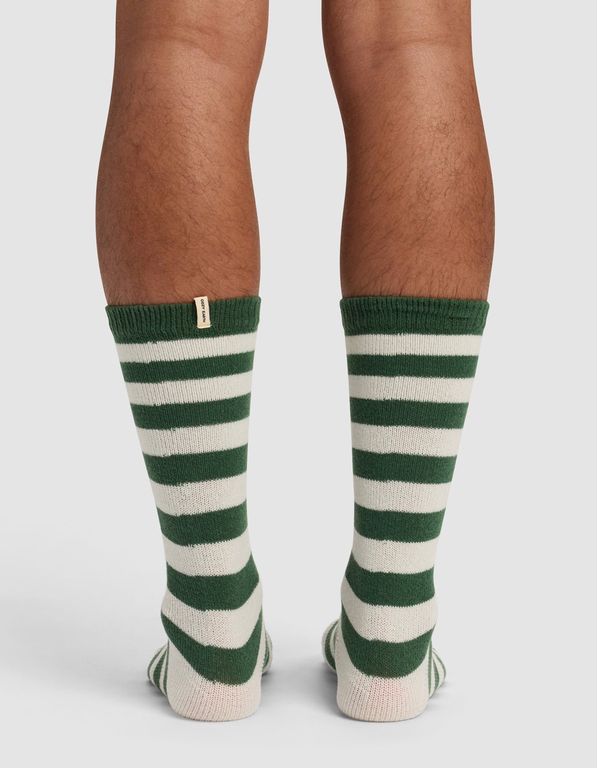 Back view of a person wearing Cozy Earth’s The Striped Plush Lounge Sock in green and white stripes, featuring a small beige tag near the top. The socks are shown on calves, standing on a light surface.