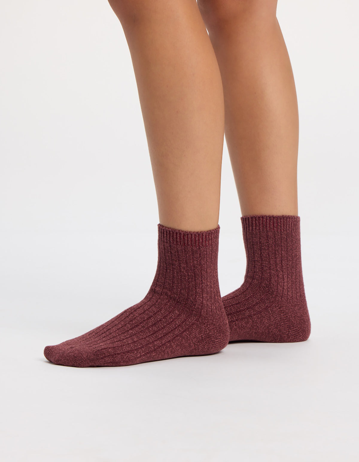 A person wearing maroon socks from the Cozy Earth Plush Lounge Quarter Sock 3-Pack stands on a white surface, with only their lower legs and feet visible.