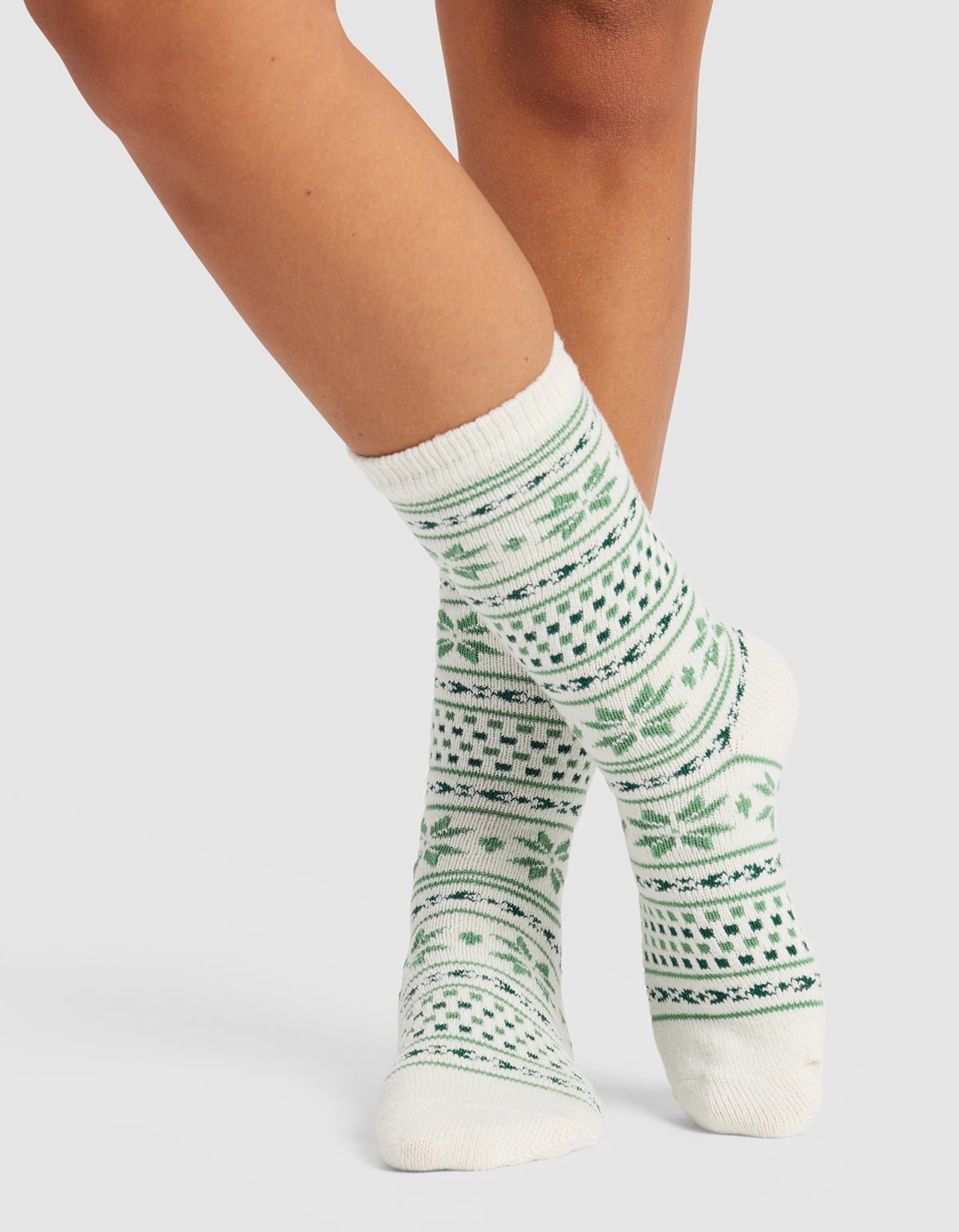 A person models Cozy Earth’s The Snowflake Plush Lounge Sock—white crew socks featuring green geometric and leaf designs—standing with one foot in front of the other against a plain background.