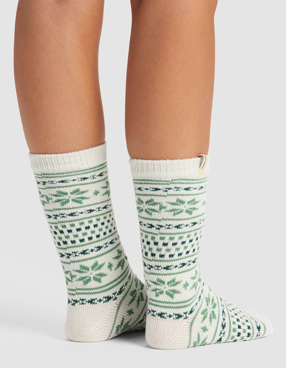 A person wears The Snowflake Plush Lounge Sock by Cozy Earth, featuring cream with green and dark green geometric and leaf designs, standing on a white surface with the back and side of their calves and feet visible.