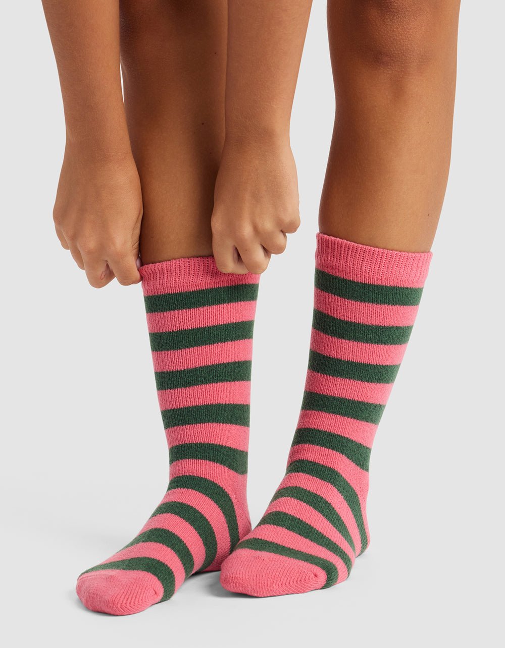 A person stands on a white surface, pulling up The Striped Plush Lounge Sock by Cozy Earth, featuring pink and dark green horizontal stripes. Only their legs and hands are visible.|Color:Hibiscus Stripe