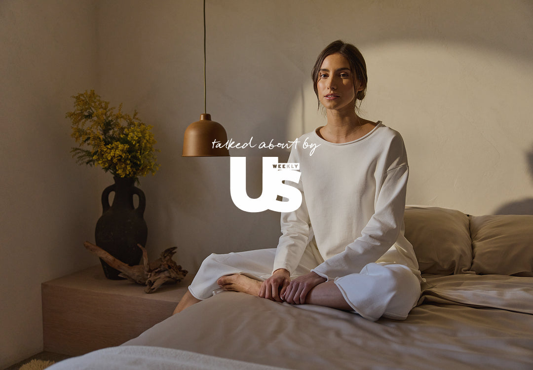 Woman sitting on a bed in a softly lit room with 'talked about by US Weekly' logo overlay.