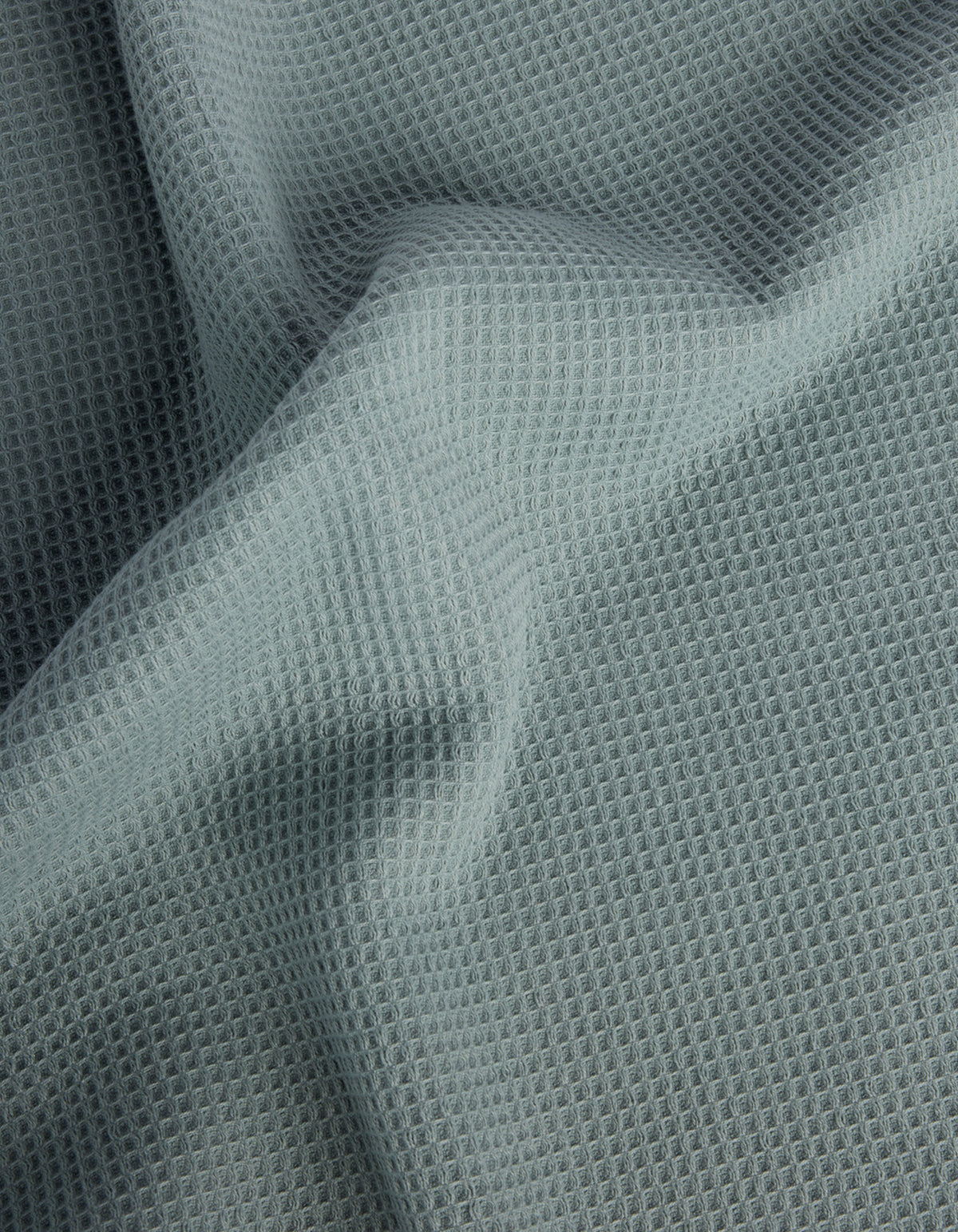 Cozy Earth’s Waffle Bath Sheets feature light gray fabric with a waffle-like texture and a grid of small, raised squares. The soft material drapes gently, creating subtle folds and shadows on the surface. 