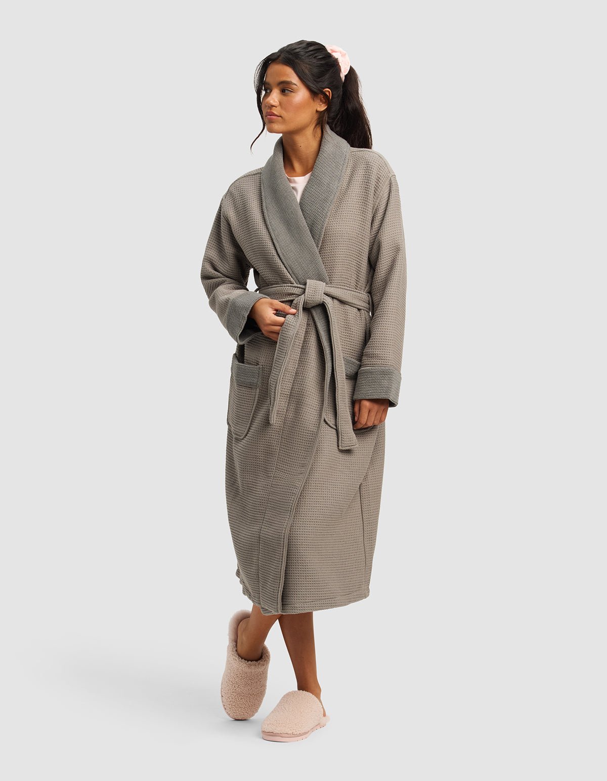 A woman with dark hair tied back wears the Cozy Earth Waffle Bath Robe—grey, textured, with large pockets and a tie belt—and pink fuzzy slippers, standing hands-in-pockets against a plain light background.|Color:Charcoal