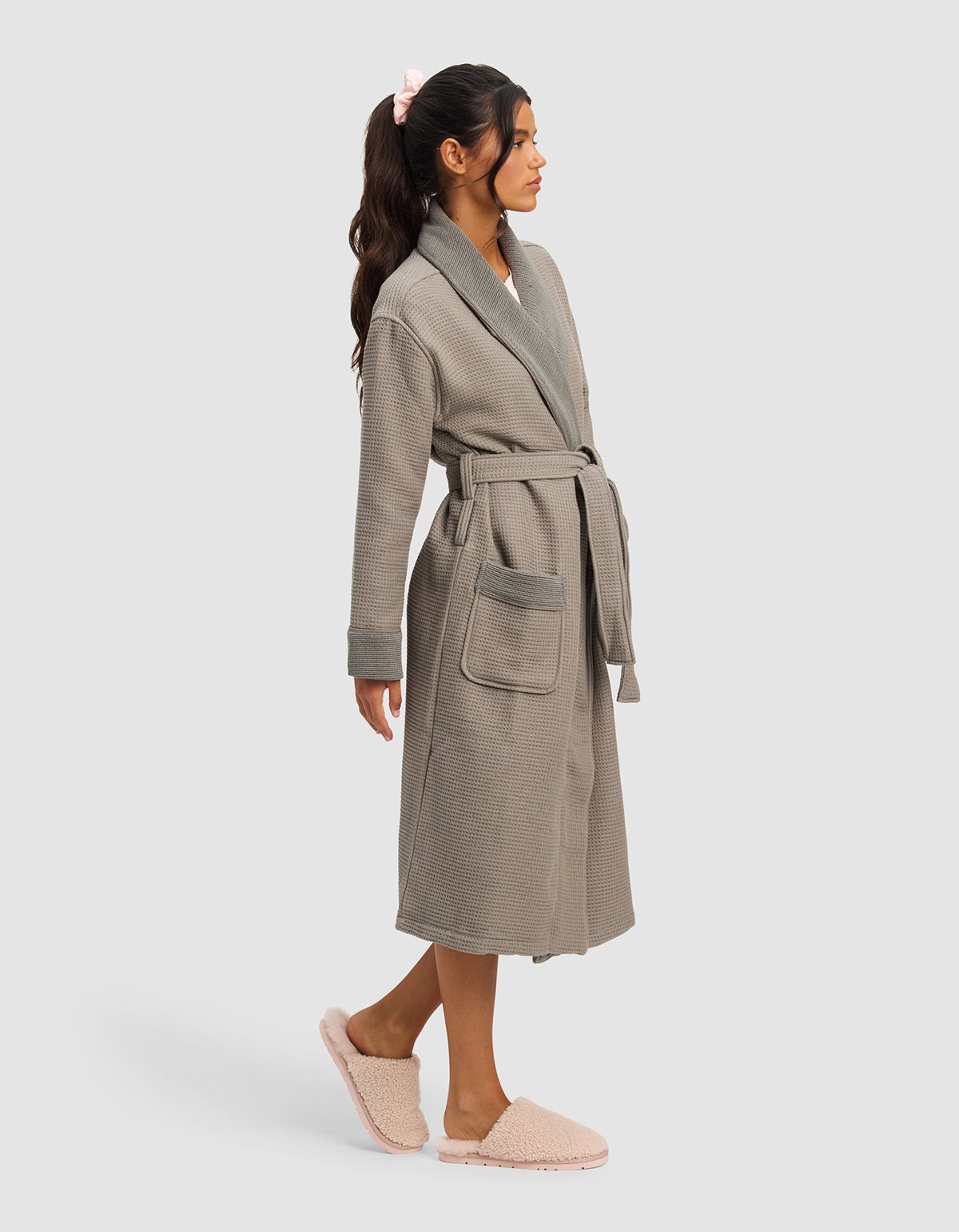 A woman with long dark hair in a ponytail wears the Cozy Earth Waffle Bath Robe in light grey, featuring large pockets and a belt, along with pink fuzzy slippers. She stands in profile against a plain light background.