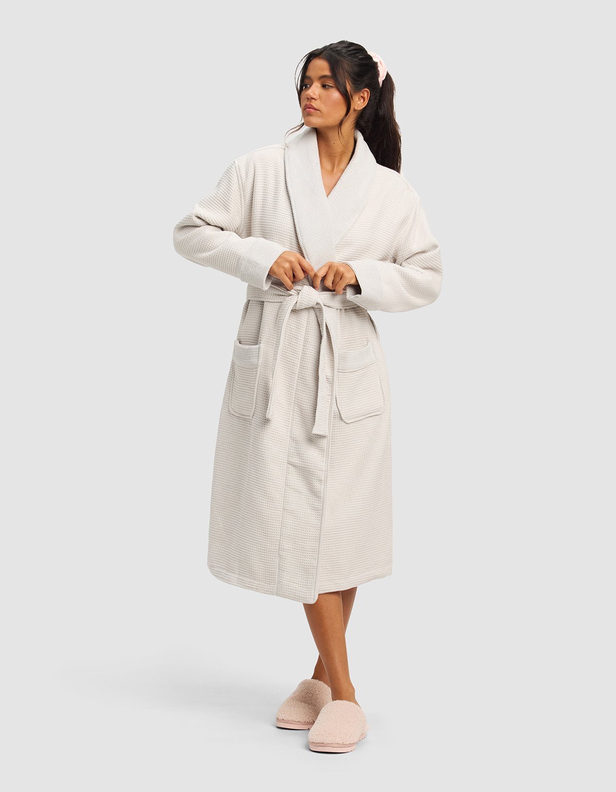 A woman models the Cozy Earth Waffle Bath Robe, featuring a belted waist and large front pockets, paired with fluffy pink slippers. She stands against a plain light background, gazing to the side.|Color:Light Grey