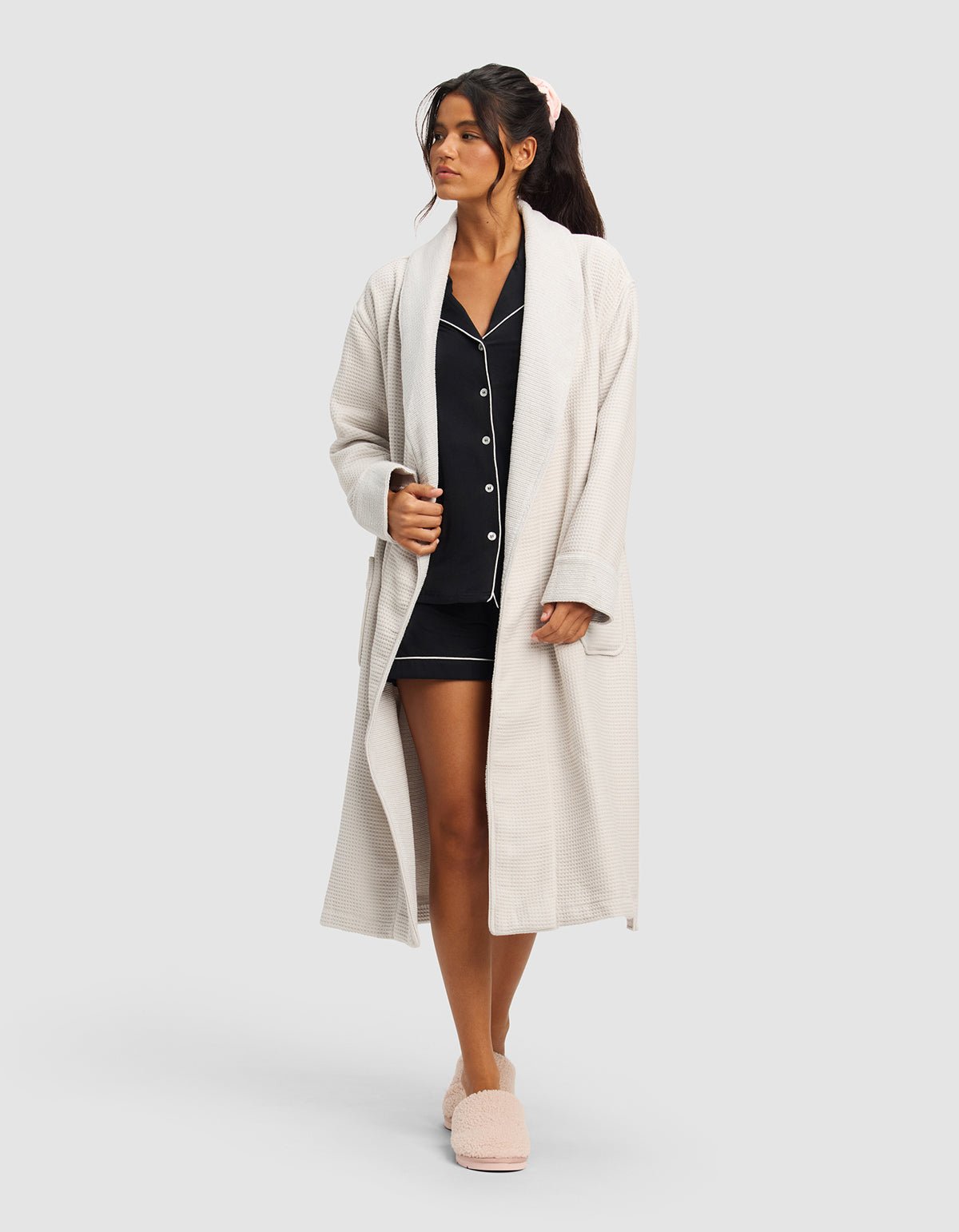 A woman wears the Cozy Earth Waffle Bath Robe over dark pajamas with white trim and matching slippers, her hair tied with a pink scrunchie as she gazes slightly to the side against a plain background.