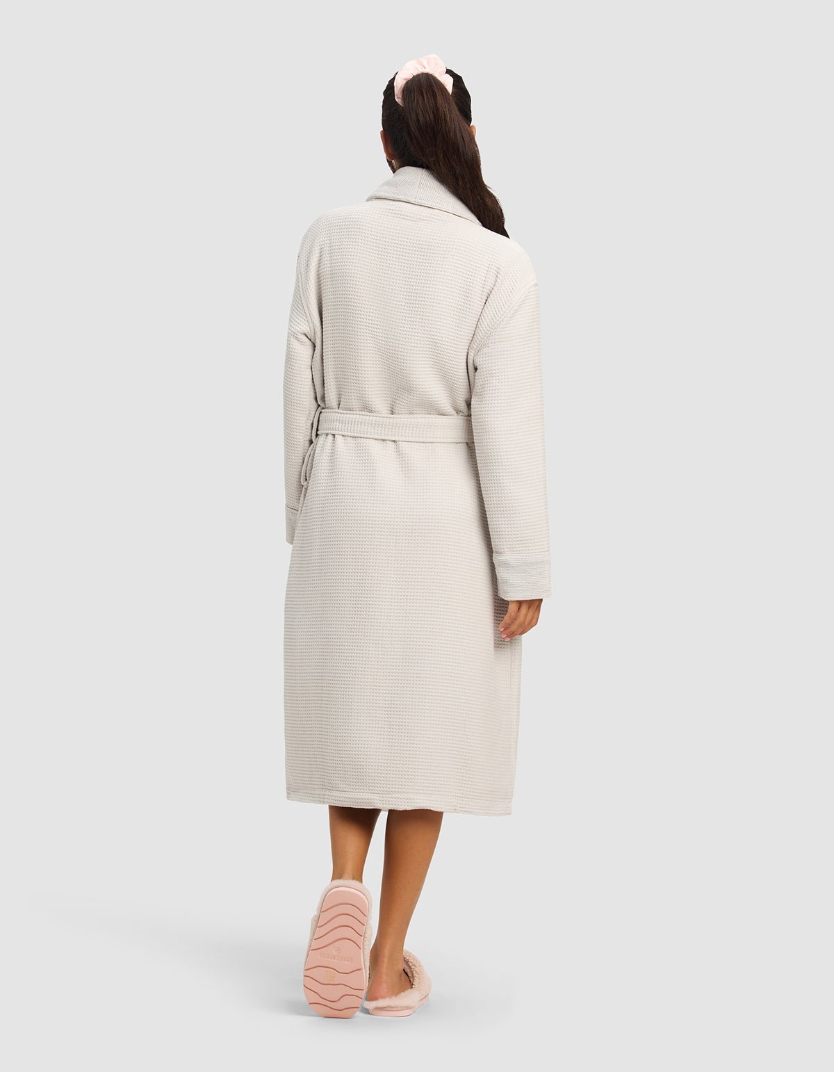 A woman seen from behind wears the Cozy Earth Waffle Bath Robe in light beige with a belt, pink slippers, and a light pink scrunchie, standing against a plain light gray background.