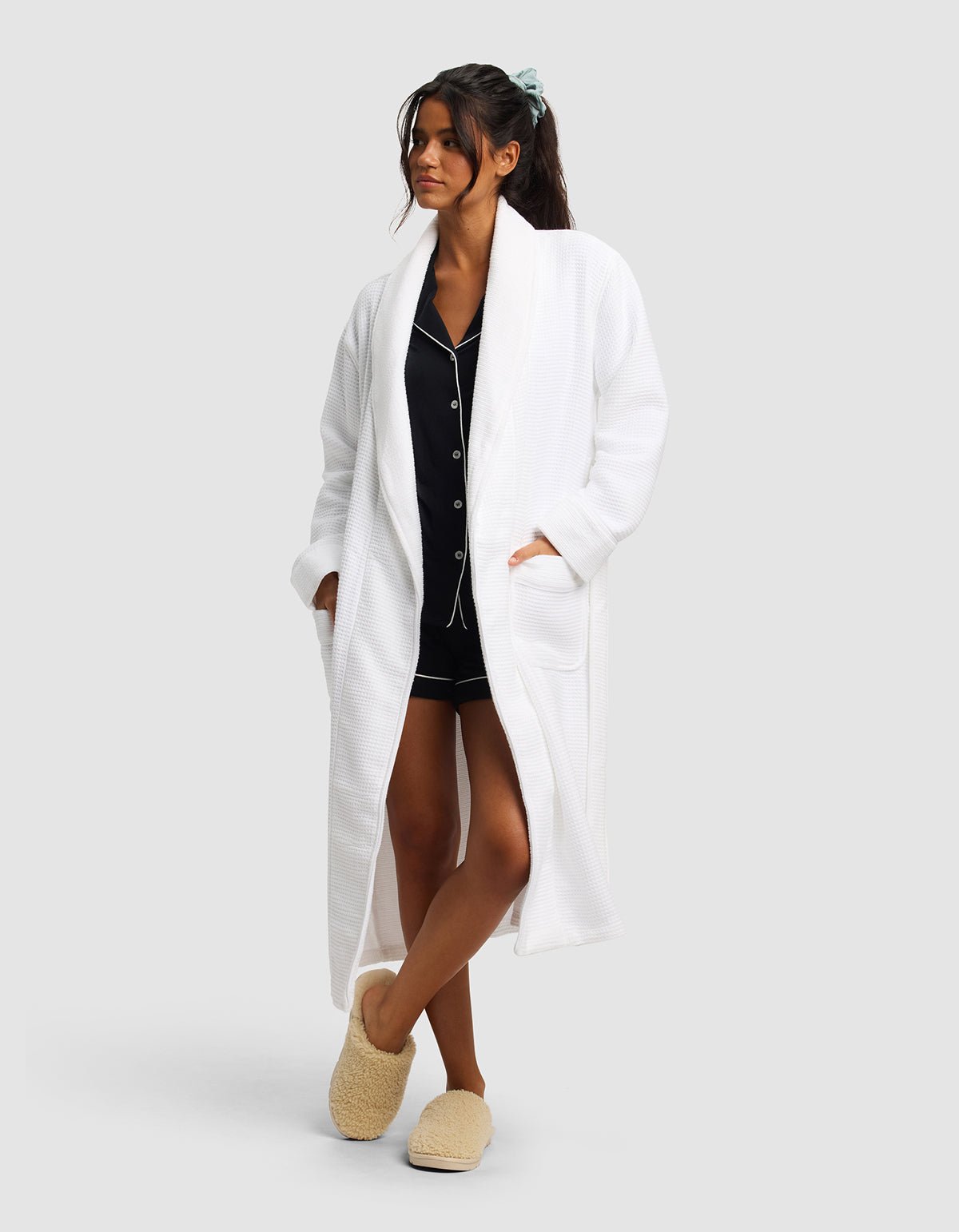A woman stands against a plain light background wearing Cozy Earth's Waffle Bath Robe over black pajamas, with her hands in the robe's pockets. She pairs the look with fluffy slippers and a blue scrunchie in her hair.