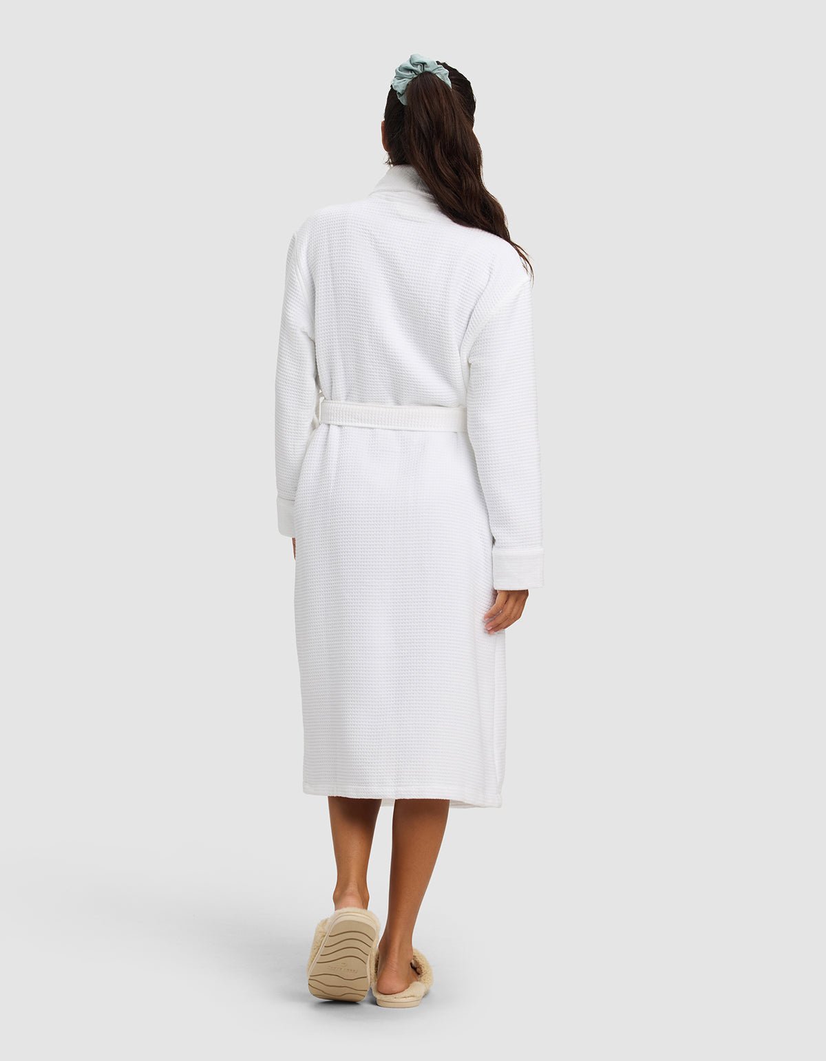 A woman with long dark hair in a green scrunchie faces away, wearing the Cozy Earth Waffle Bath Robe in white and tan slippers, standing against a plain light gray background.