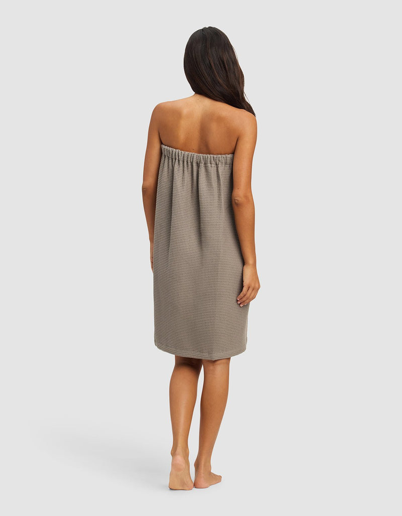 A woman with long, dark hair stands barefoot on a plain background, facing away from the camera. She wears the Cozy Earth Waffle Bath Wrap, a strapless, knee-length towel wrap in taupe with a textured pattern.