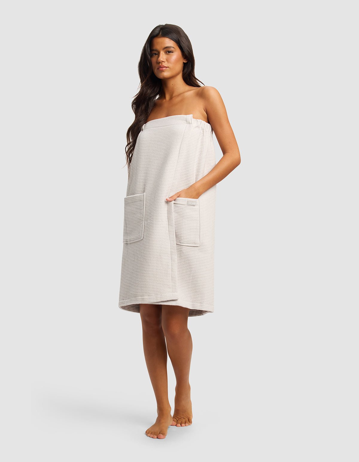 A woman with long dark hair stands barefoot wearing the Cozy Earth Waffle Bath Wrap in light beige, featuring two large front pockets and covering her from chest to knees. The background is plain and light gray.