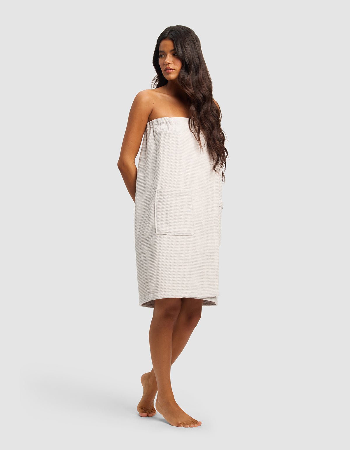 A woman with long dark hair stands barefoot, wearing the Cozy Earth Waffle Bath Wrap—a strapless, light towel wrap with large front pockets—facing slightly to the side against a plain, light background.|Color:Light Grey