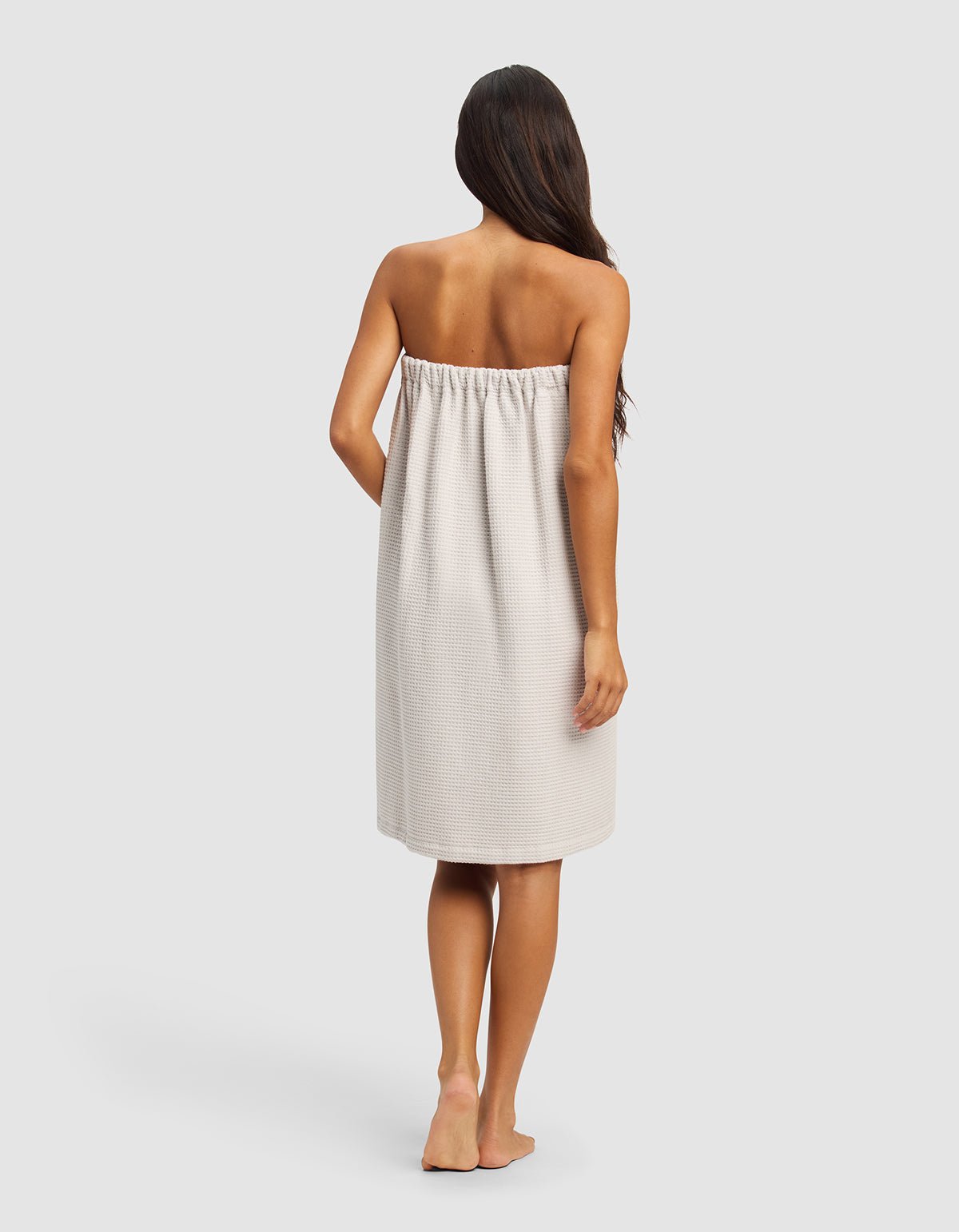 A woman with long dark hair stands barefoot, facing away, wearing the Cozy Earth Waffle Bath Wrap in light beige, covering her from chest to knees. The background is plain and light gray.