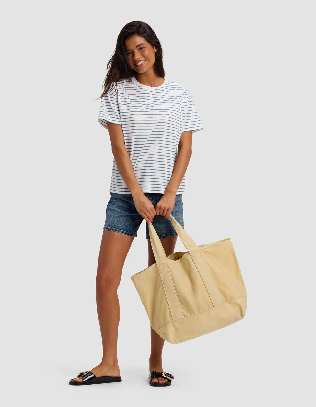 A woman stands smiling against a light gray background, holding the Cozy Earth Waxed Canvas Tote with both hands, dressed in a white and navy striped t-shirt, denim shorts, and black sandals.