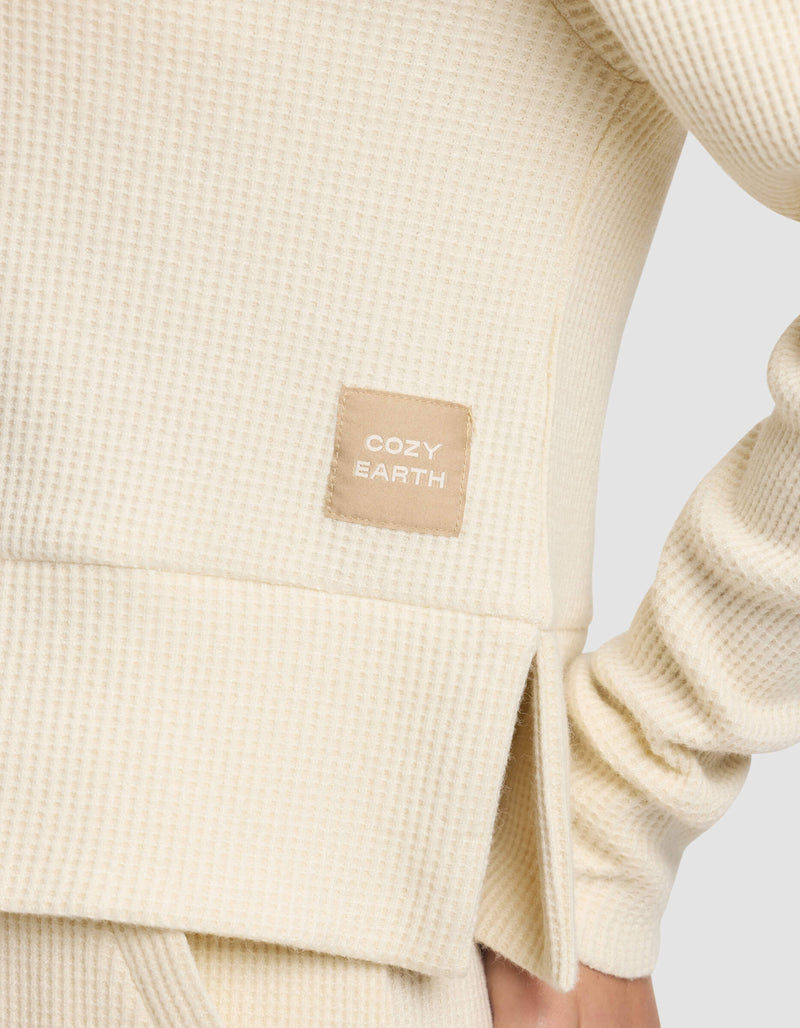 Close-up of the Women's Blake Waffle Hoodie by Cozy Earth in cream, showcasing its textured fabric and a stitched "COZY EARTH" tag. The wearer’s arm is bent at the elbow.