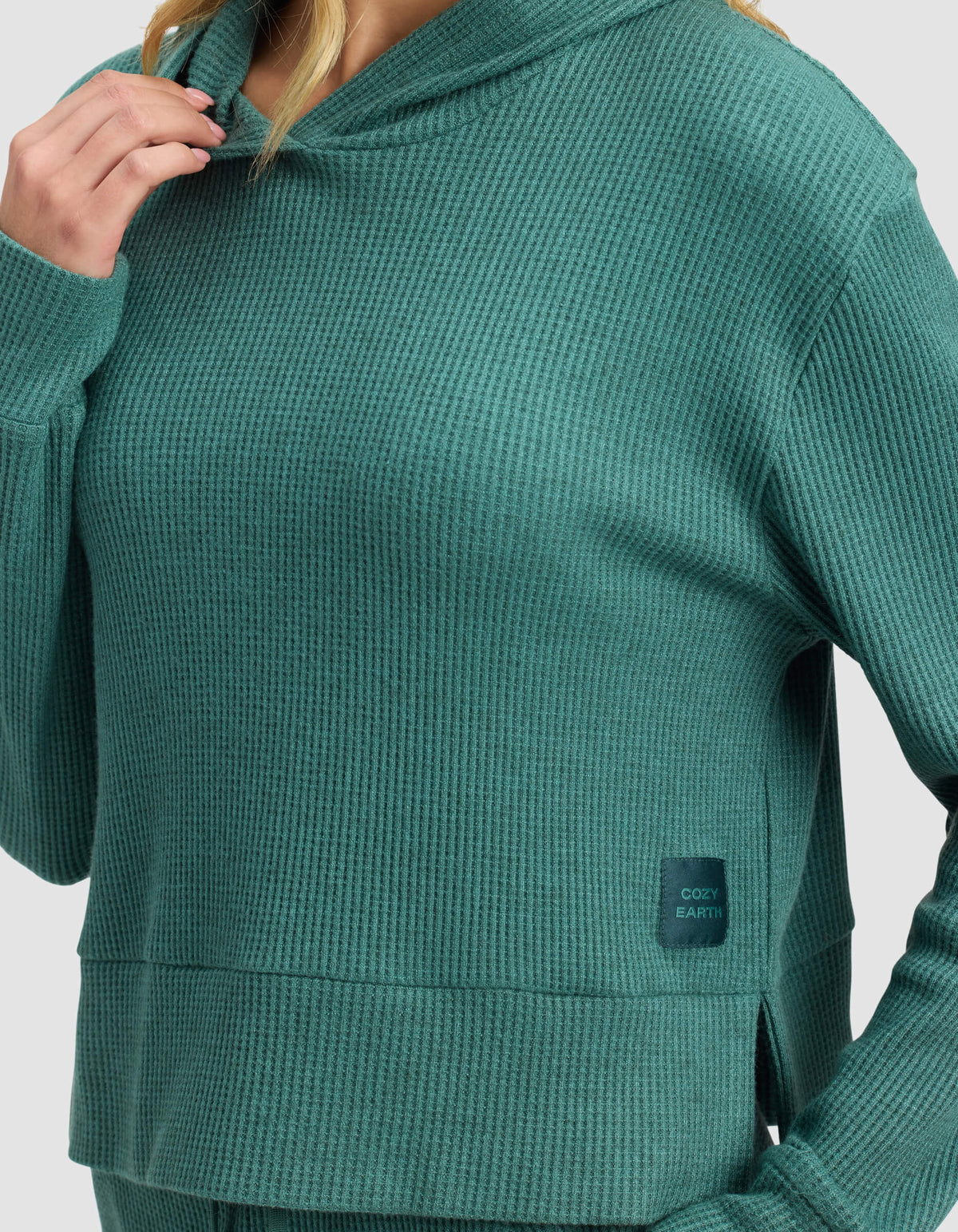 A person wears the Cozy Earth Women's Blake Waffle Hoodie in textured teal-green, featuring a "COZY EARTH" patch on the lower front. The face is partially out of view.
