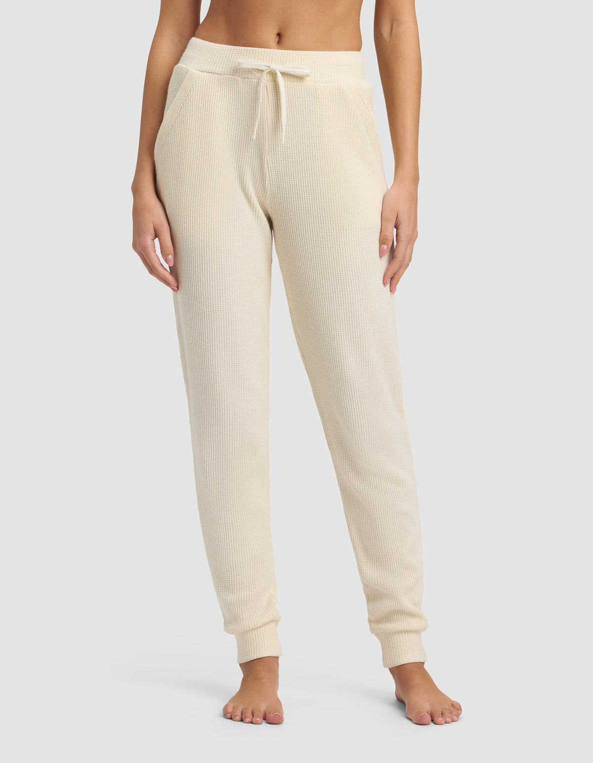 A person wears Cozy Earth's Women's Blake Waffle Jogger in cream, featuring a ribbed knit, drawstring waist, and pockets, while standing barefoot against a plain background.