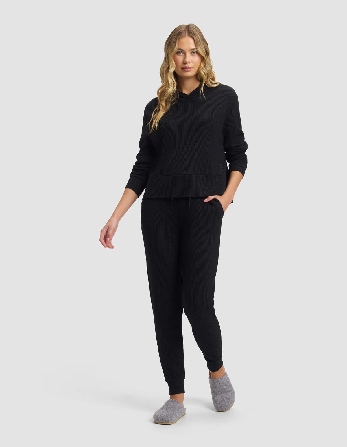 A woman with long blonde hair wears the Cozy Earth Women's Blake Waffle Jogger set with gray slippers, standing and looking forward against a plain light gray background.