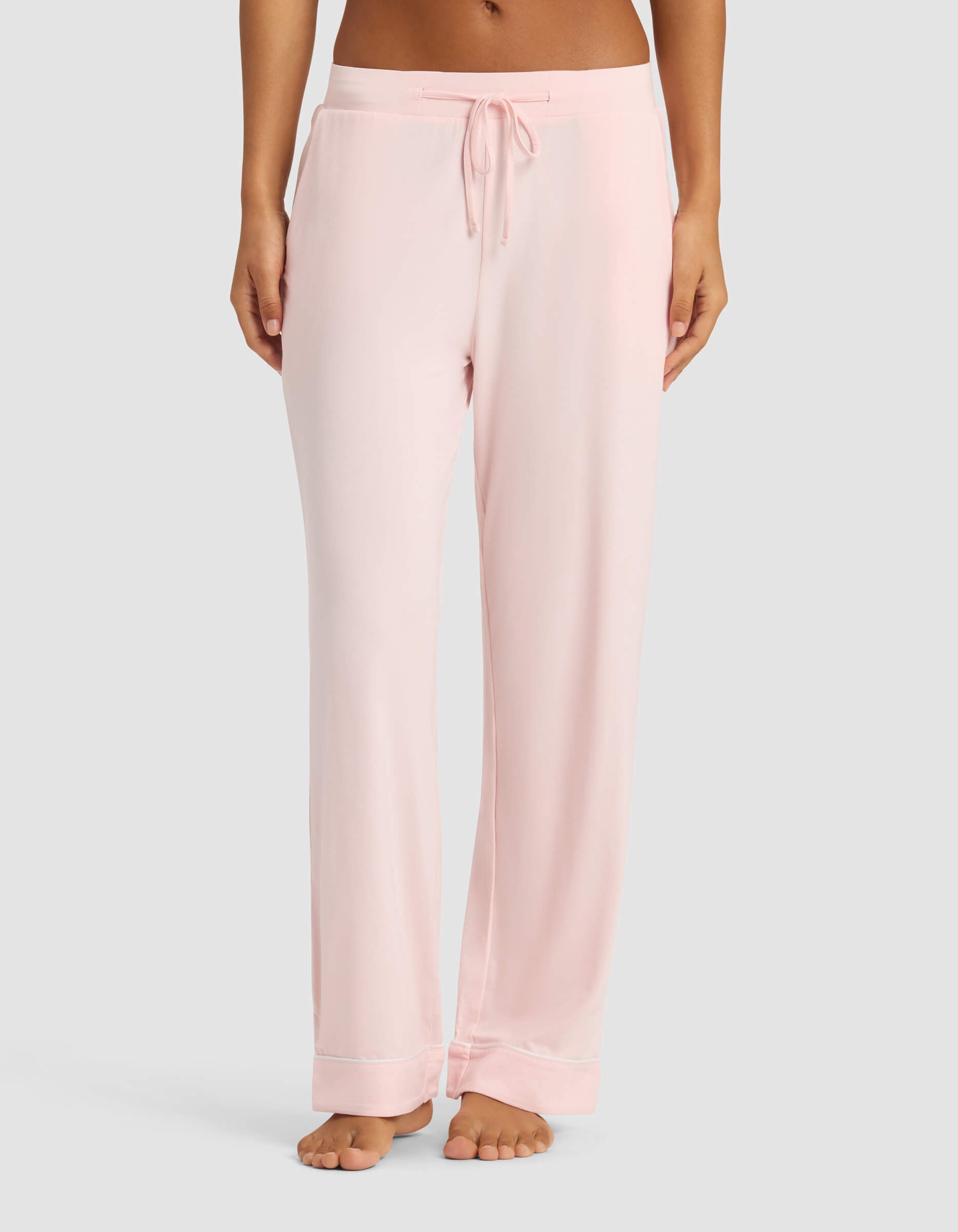 A person wears Cozy Earth Women’s Bamboo Stretch-Knit Pajama Pant in light pink, featuring a drawstring waist and pockets. Only the lower torso and legs are seen, standing barefoot against a plain gray background.|Color:Blossom