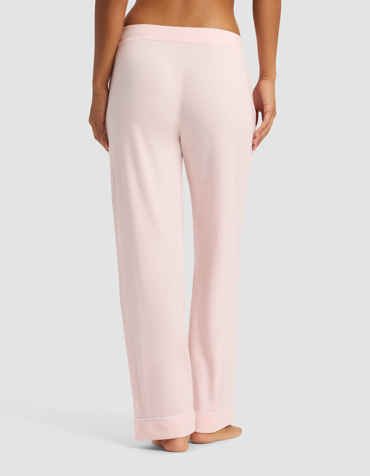 A person is standing barefoot on a neutral background, shown from the back, wearing Cozy Earth Women's Bamboo Stretch-Knit Pajama Pants in light pink with a wide waistband.