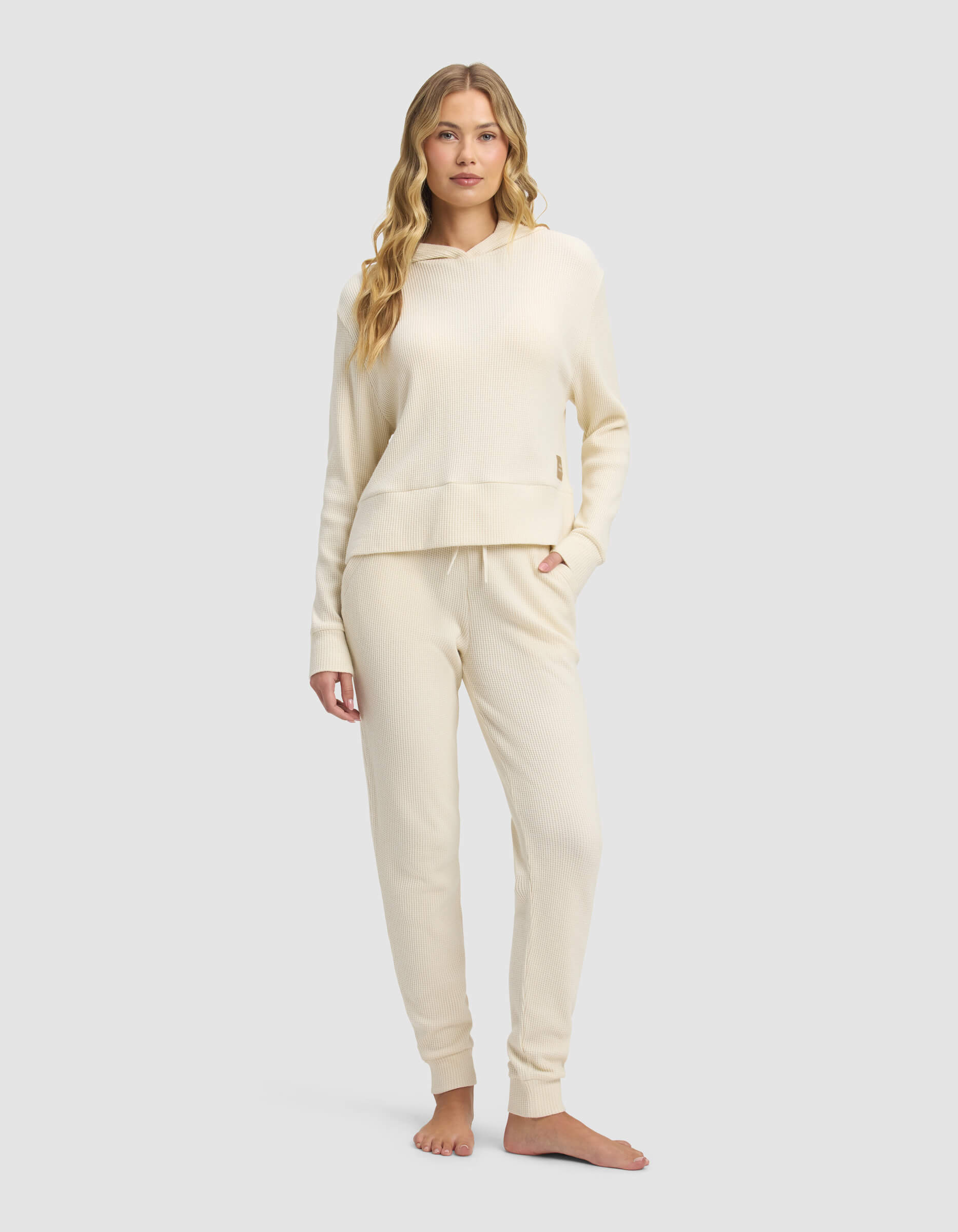 A woman stands barefoot against a light gray background, wearing the Cozy Earth Women's Blake Waffle Hoodie & Jogger Set in cream. She faces forward with one hand in her pocket.|Color:Coconut