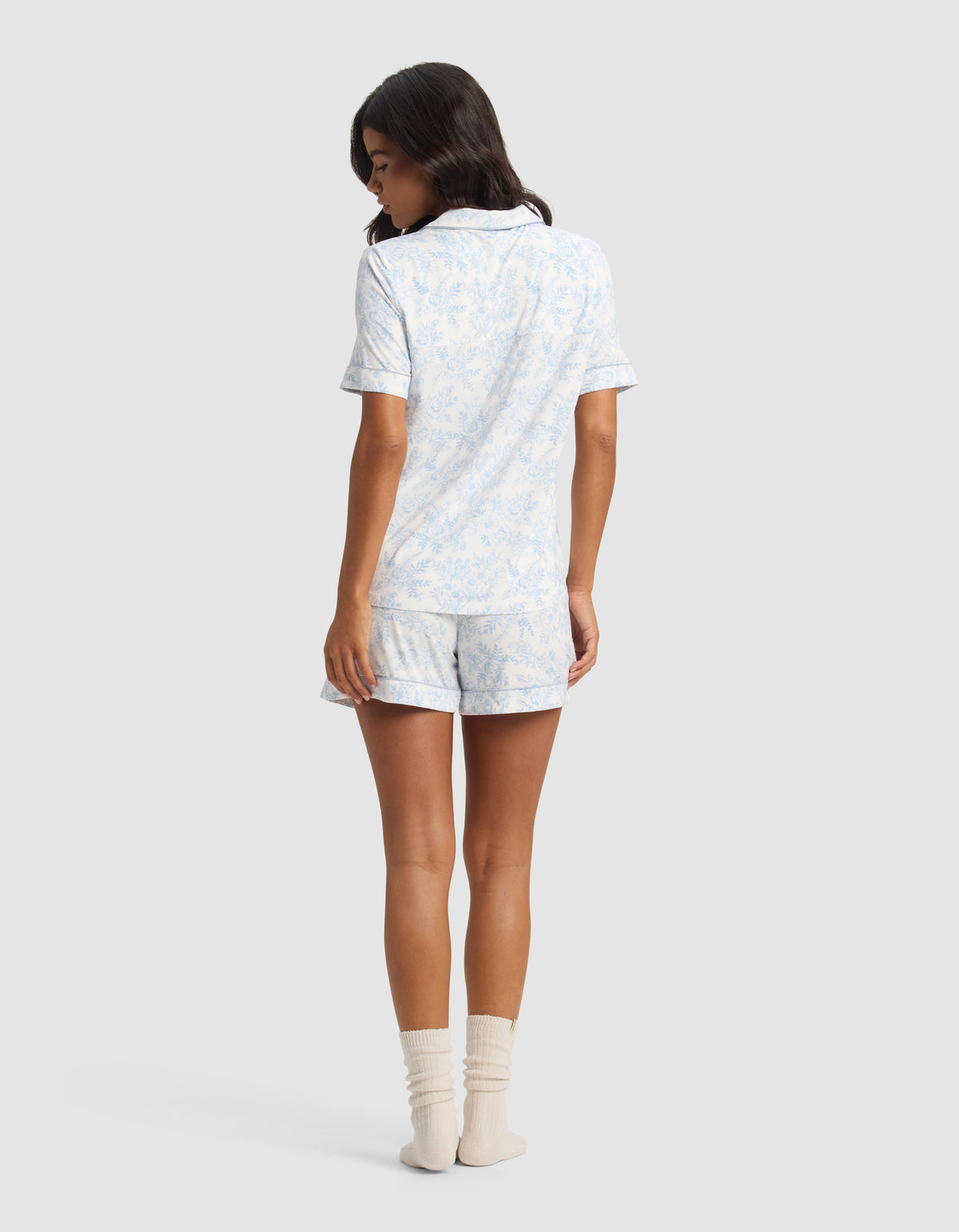 A woman with medium-length dark hair wears the Cozy Earth Women's Bamboo Stretch-Knit Short Sleeve Pajama Set in light blue and white, paired with cream socks, facing away against a plain light background.