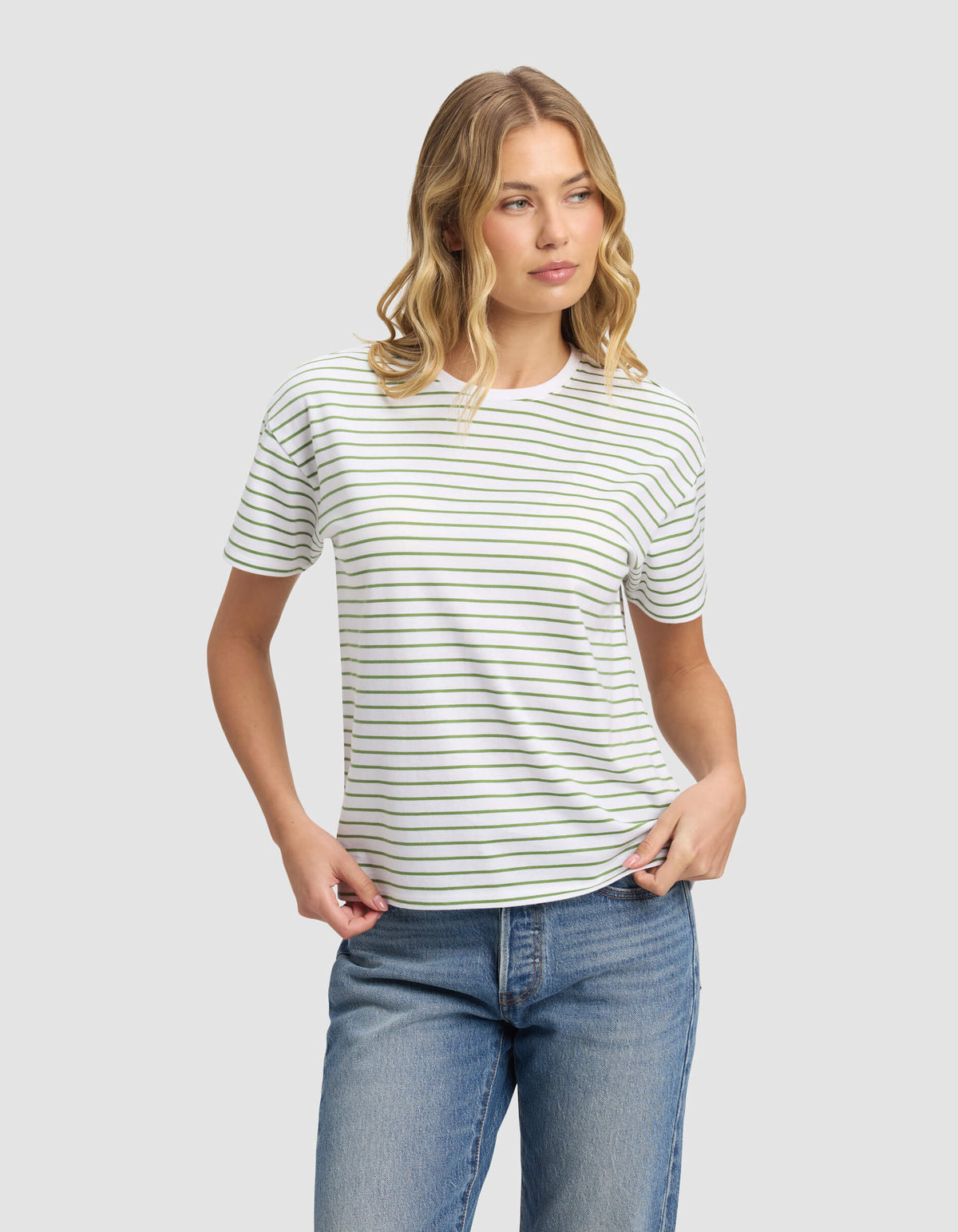 A woman with wavy blonde hair models the Cozy Earth Women's All Day Tee, a white short-sleeve shirt with thin black stripes, paired with blue jeans. She stands against a plain light gray background, gazing to the side in a relaxed mood.
