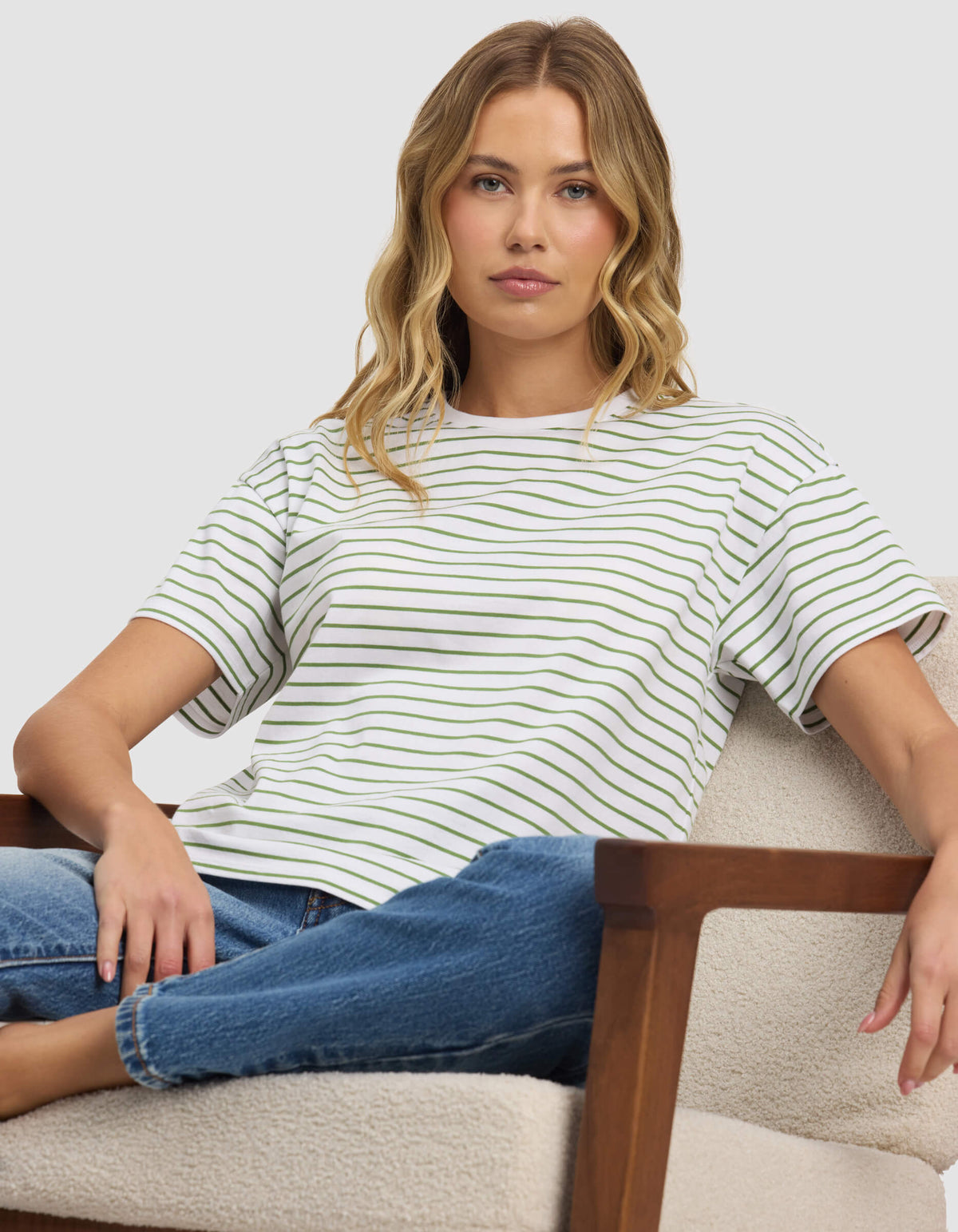 A woman with wavy blonde hair sits on a light-colored armchair, wearing Cozy Earth's Women's All Day Tee and blue jeans, looking directly at the camera with a neutral expression.
