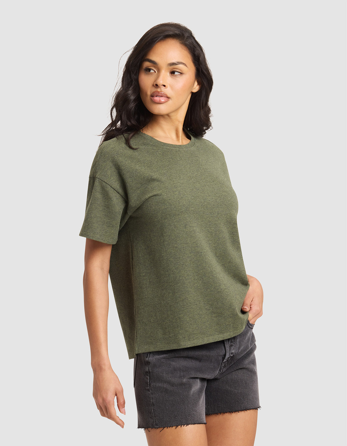 A woman with long dark hair models the Cozy Earth Women's All Day Tee in olive green, paired with black denim shorts. She stands against a plain light gray background, looking slightly left with one hand in her pocket.