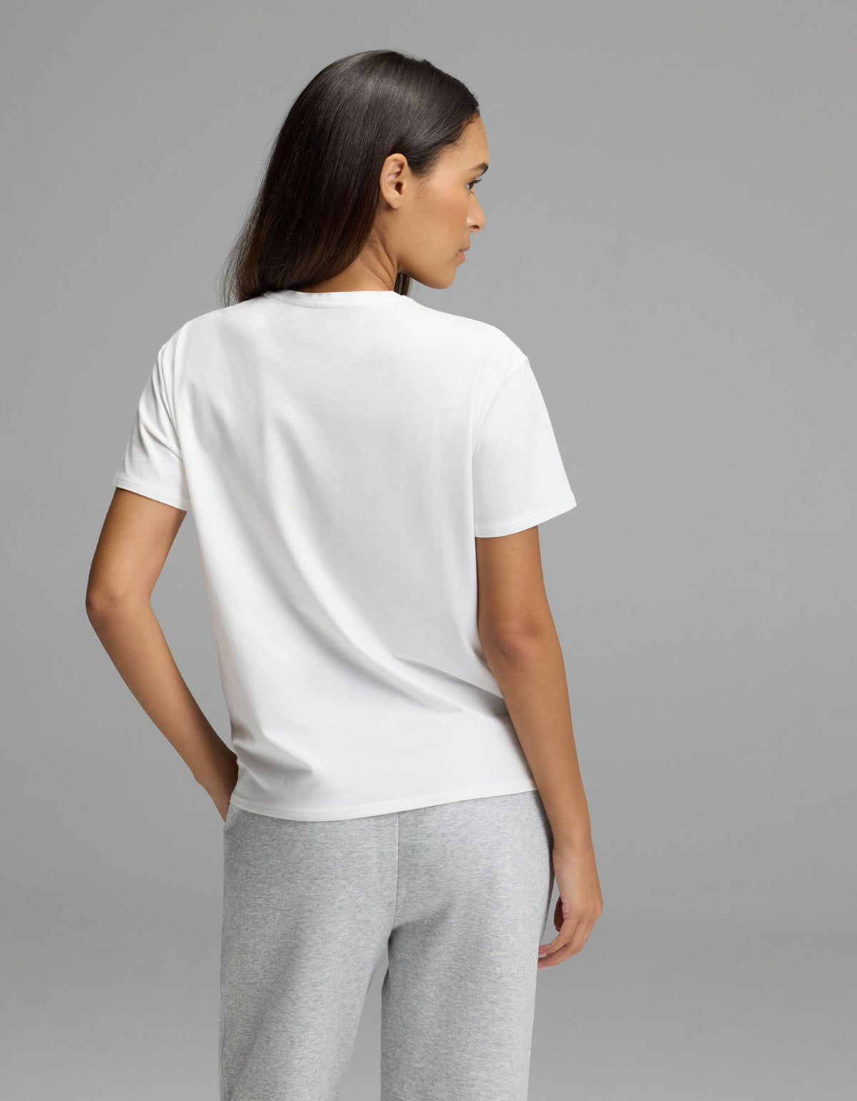 A woman with long dark hair wears the Cozy Earth Women’s All Day Tee in white and gray pants, standing against a plain gray background and looking to the side.