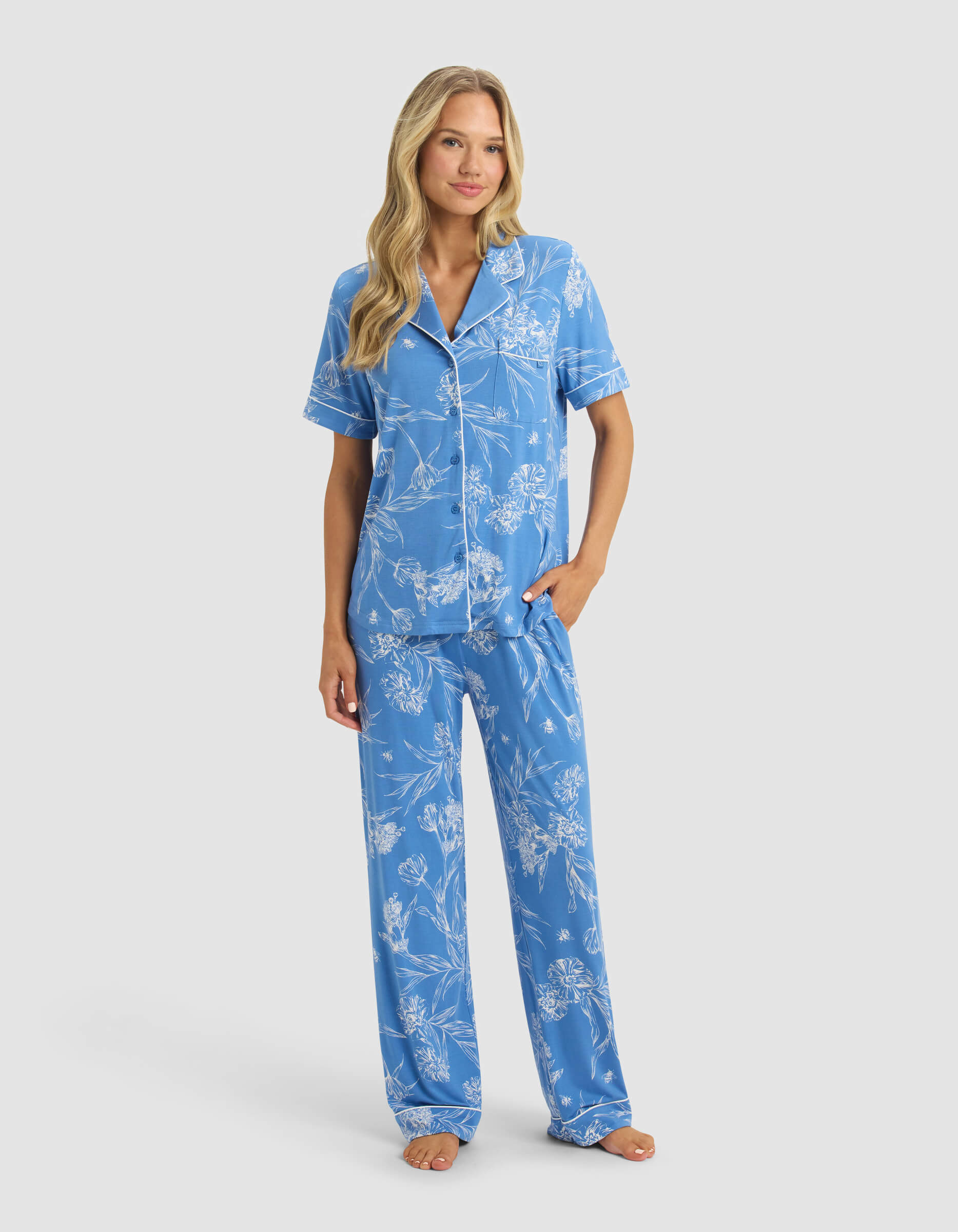 A woman with long blonde hair wears the Cozy Earth Women's Bamboo Stretch-Knit Short Sleeve & Pant Pajama Set in blue with white floral patterns. She stands barefoot against a plain light background.|Color:Garden Toile in Sailor