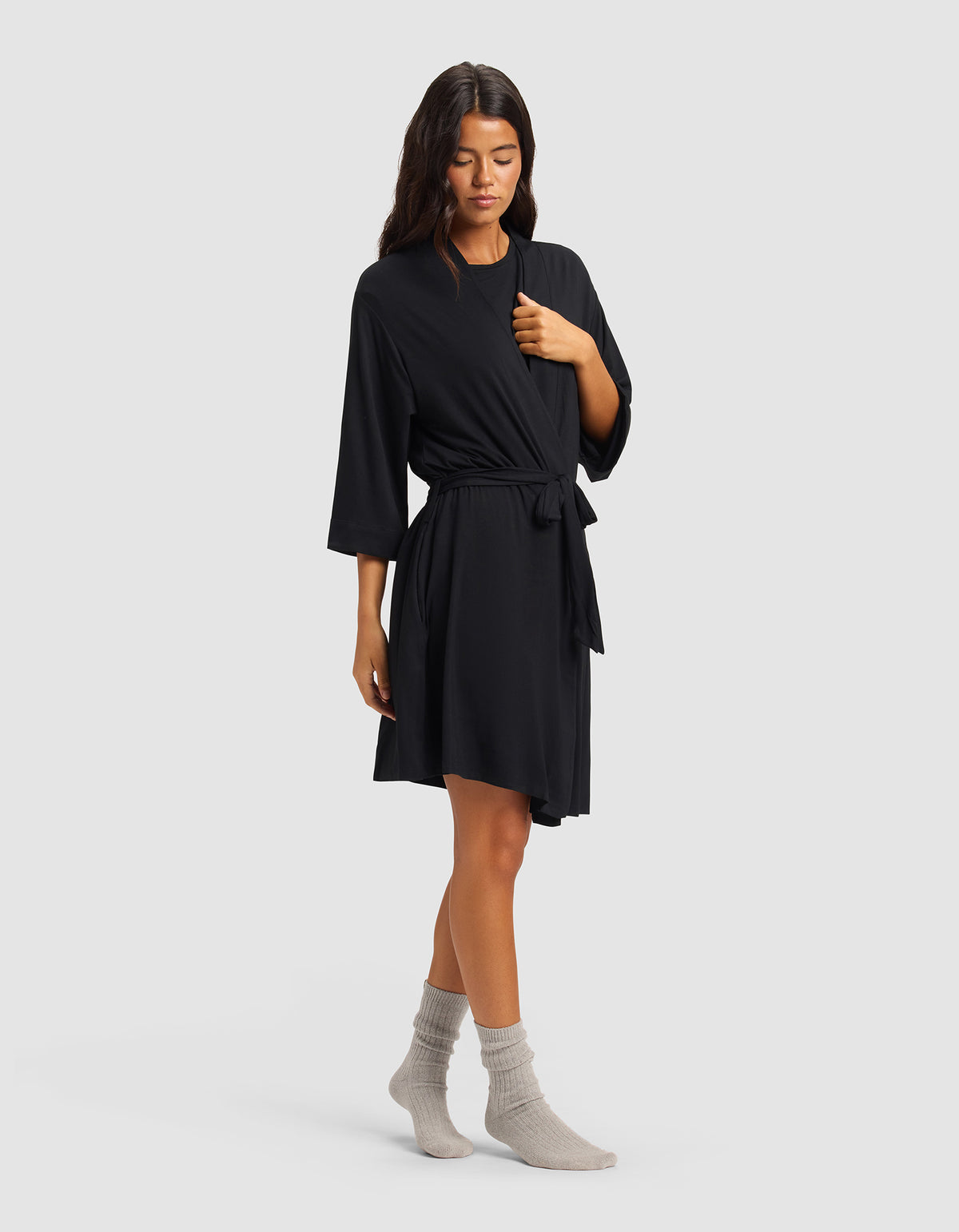 A woman with long dark hair stands against a plain background wearing the Cozy Earth Women's Bamboo Stretch-Knit Kimono Robe in black, featuring three-quarter sleeves and a tie waist, paired with light gray socks. She has a relaxed posture.
