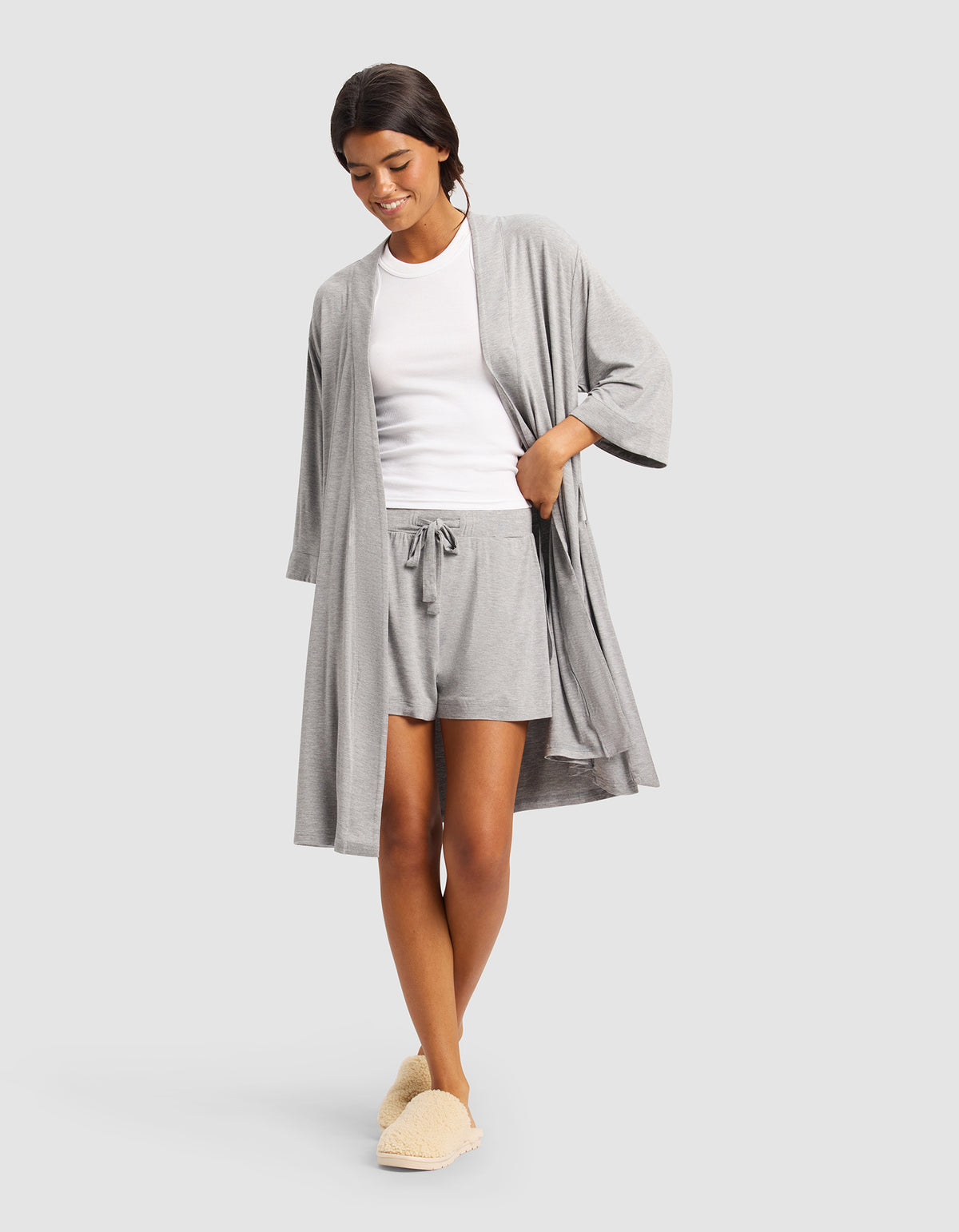 A woman wearing Cozy Earth’s Women's Bamboo Stretch-Knit Kimono Robe in grey, paired with a white T-shirt, matching grey shorts, and cream slippers, smiles with one hand in her pocket against a plain light gray background.