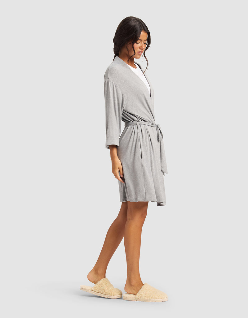 A woman stands sideways against a plain light background, relaxed in Cozy Earth’s Women's Bamboo Stretch-Knit Kimono Robe in light gray with a waist tie, paired with beige slippers. Her loose waves add to the comfortable vibe.