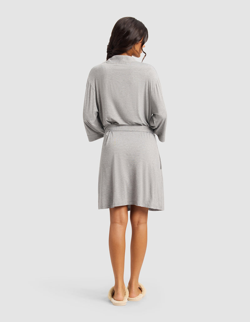 A woman with long dark hair, seen from the back, wears a Cozy Earth Women's Bamboo Stretch-Knit Kimono Robe in light gray with a belt and cream slippers, standing against a plain light background.